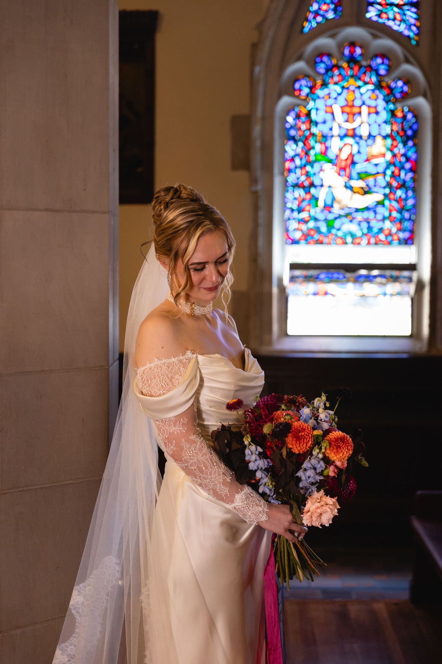 A bride in an off-shoulder wedding dress with a veil poses near a stained glass church window with a colorful bouquet.