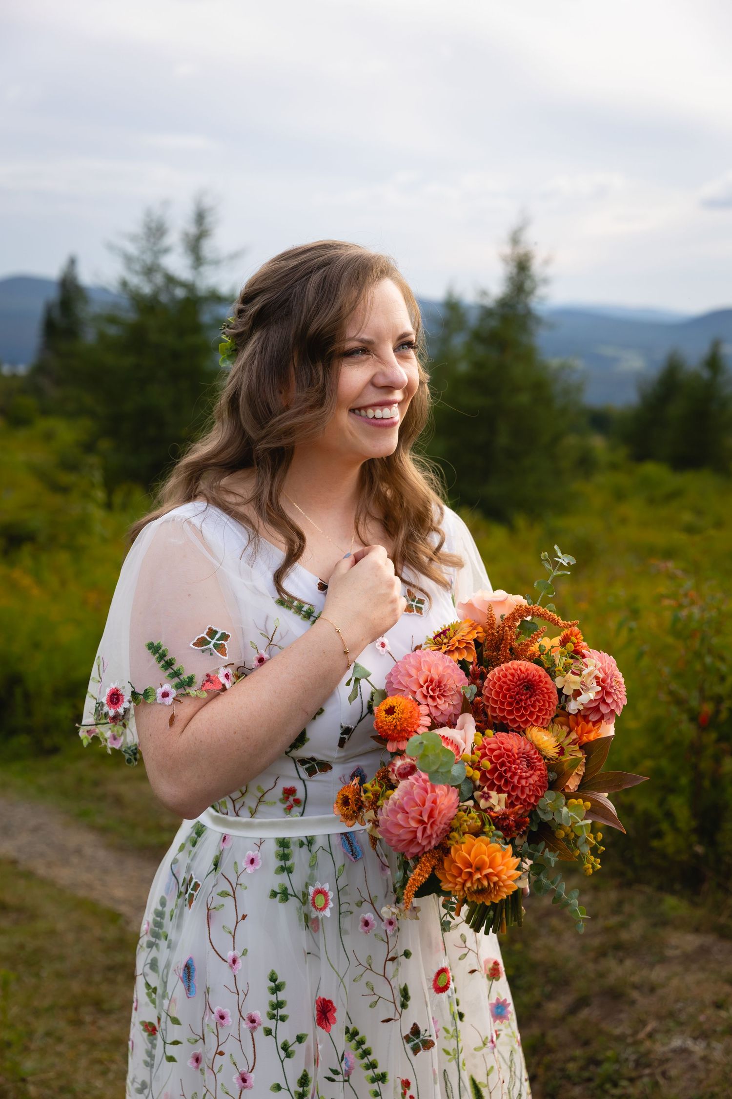 Woman in floral dress holding vibrant orange and coral dahlias while standing in mountain meadow.