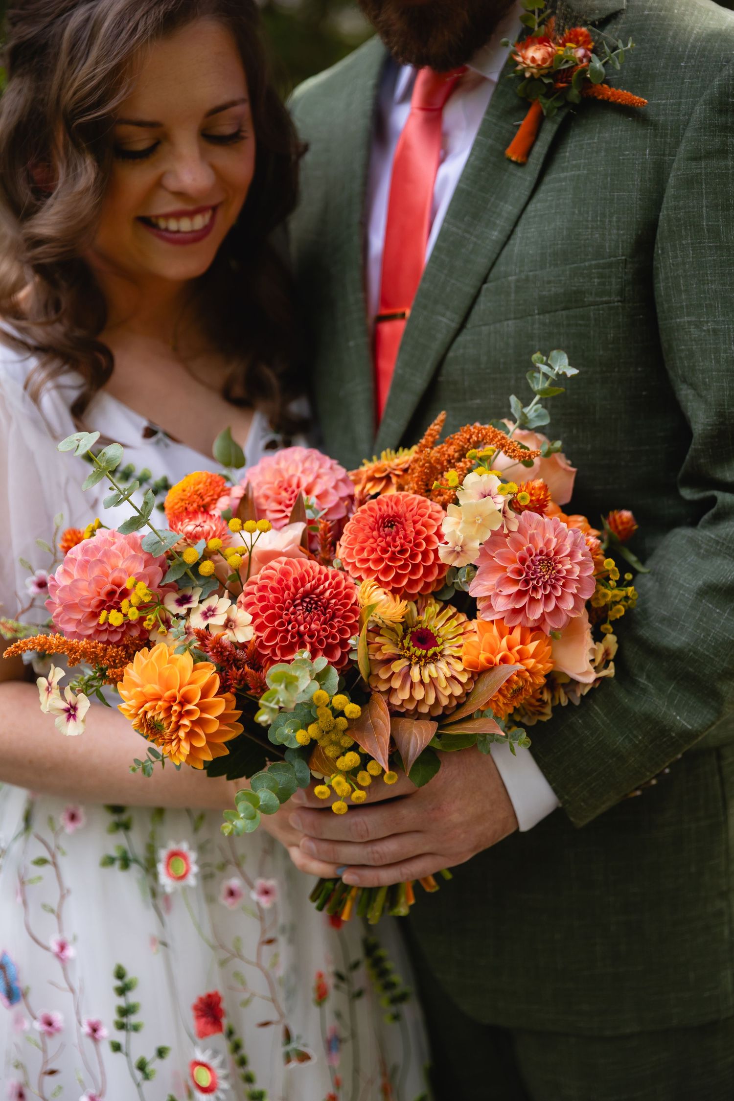 A wedding couple holds vibrant orange and coral dahlias in their autumn-themed floral bouquet against formal attire.
