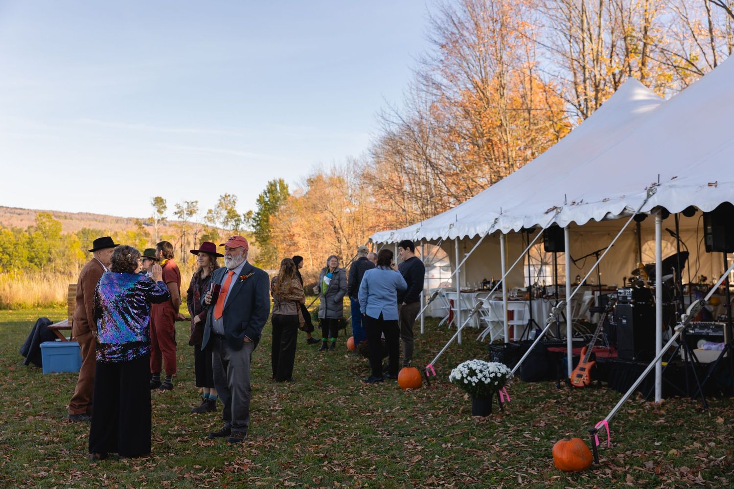 A group of people gather outdoors at a fall festival with white tents and pumpkins visible in the autumn landscape.
