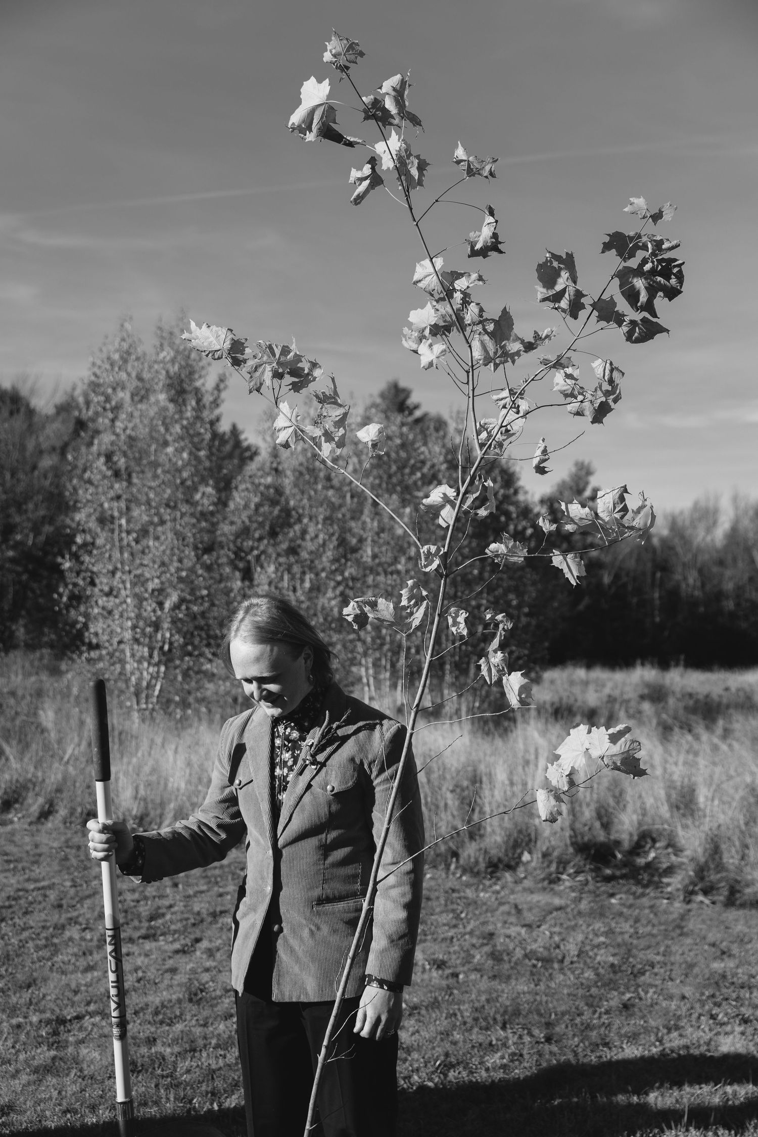 Black and white photograph showing a figure standing in a field next to young trees on an autumn day.