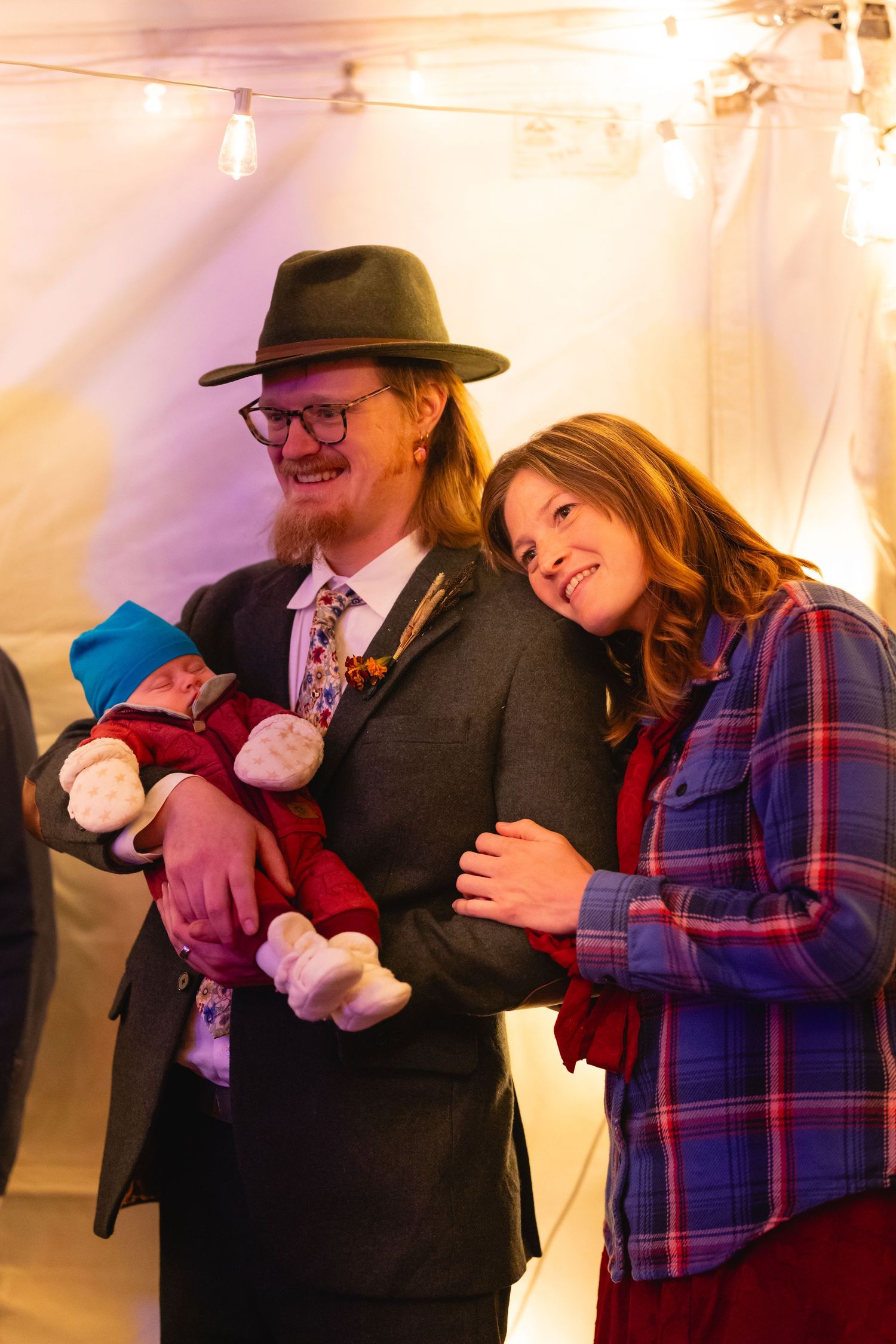 A couple in festive attire share a tender moment while holding a small baby at an indoor event with string lights.