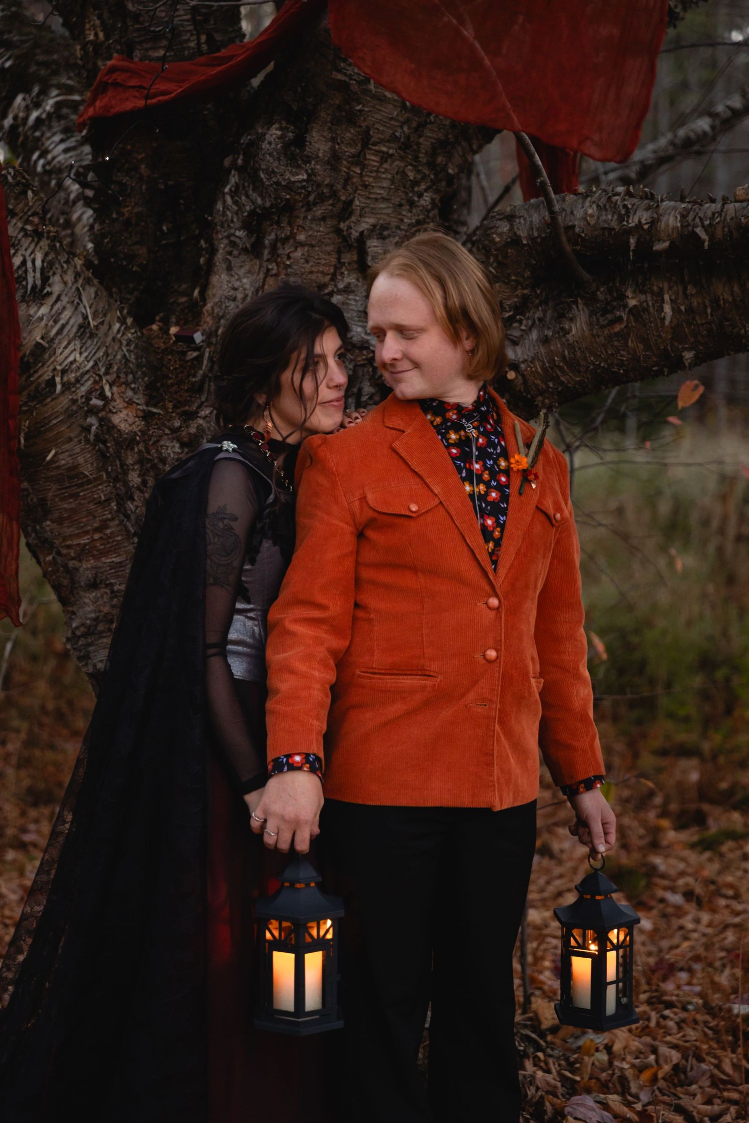 Two people in autumn attire holding lanterns walk together through fallen leaves in a dark wooded setting.