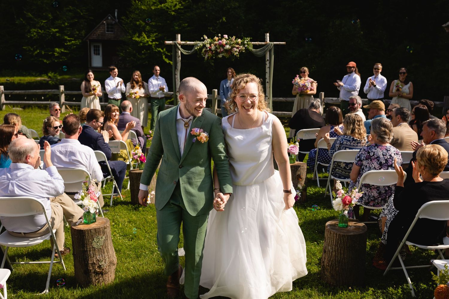 Guests enjoy a sunny outdoor wedding reception with tables set up on a grassy lawn surrounded by trees.