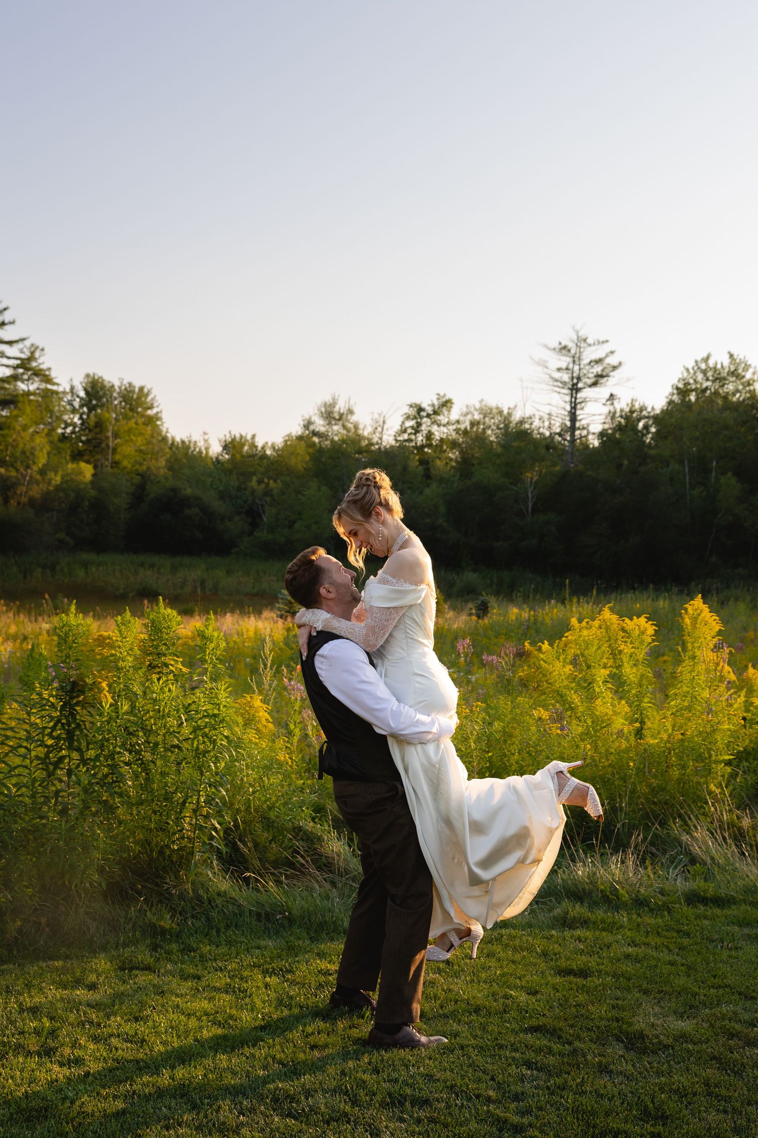 A romantic wedding couple embraces in a golden meadow at sunset, with the groom lifting the bride in her flowing white dress.