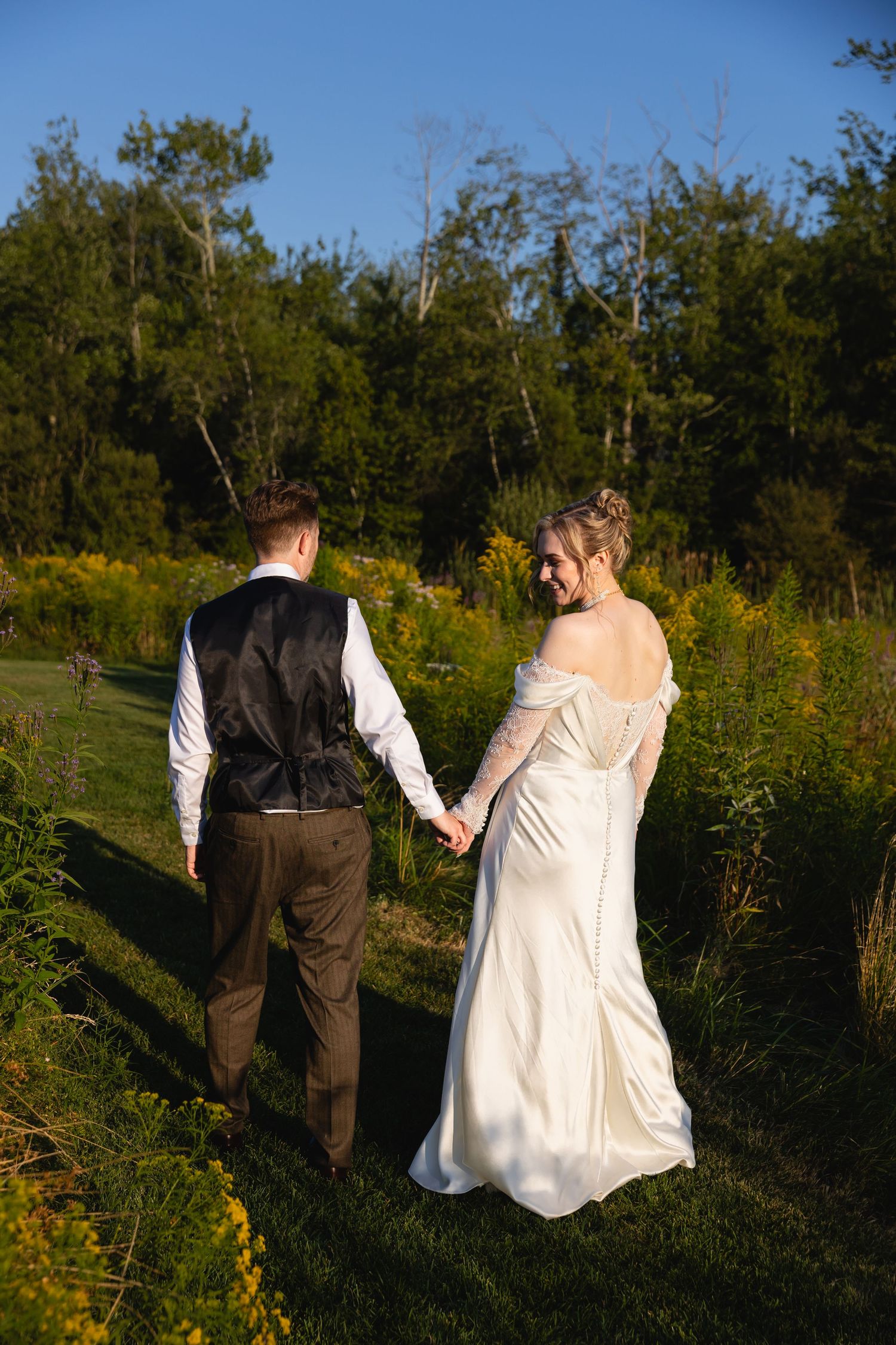 A couple walks through a sunlit meadow with tall grass and wildflowers during golden hour.