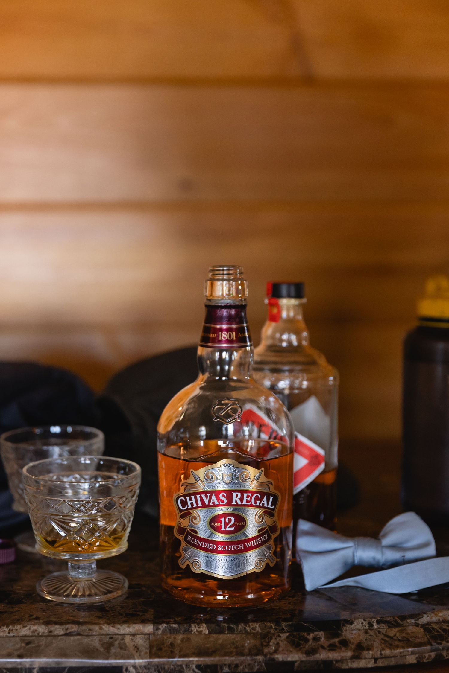 A crystal glass and bottle of Chivas Regal whisky sit on a rustic wooden surface in warm lighting.