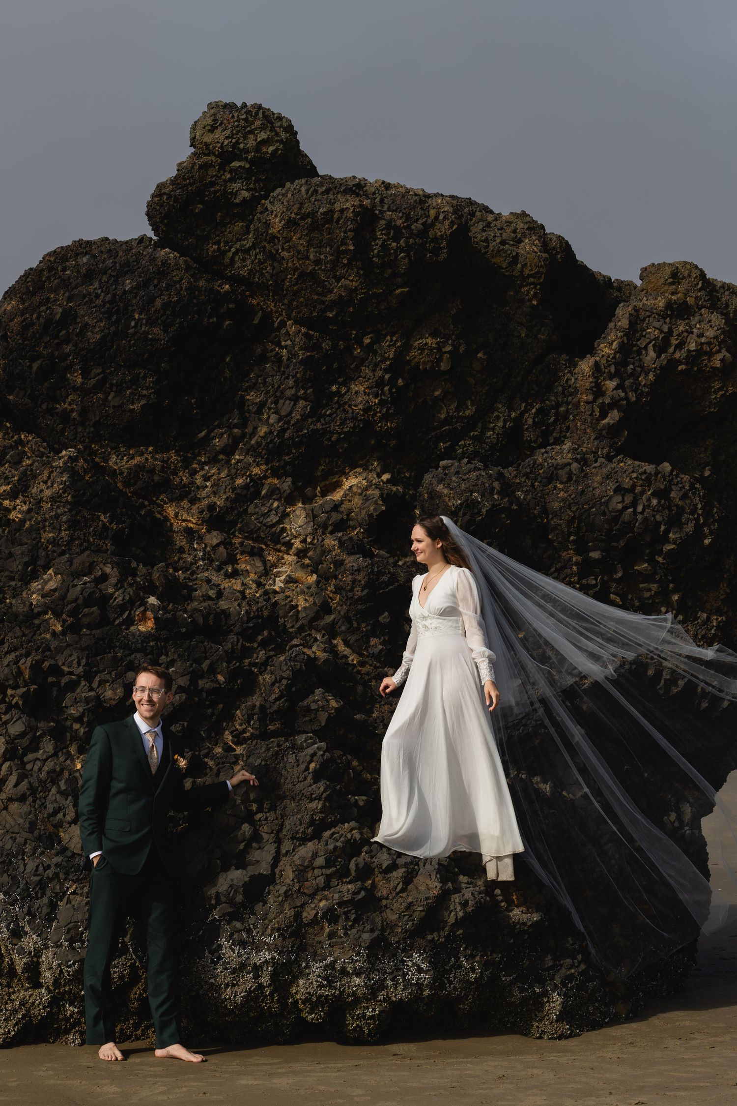 A wedding couple poses dramatically against dark volcanic rocks on a beach as a long white veil flows in the wind.