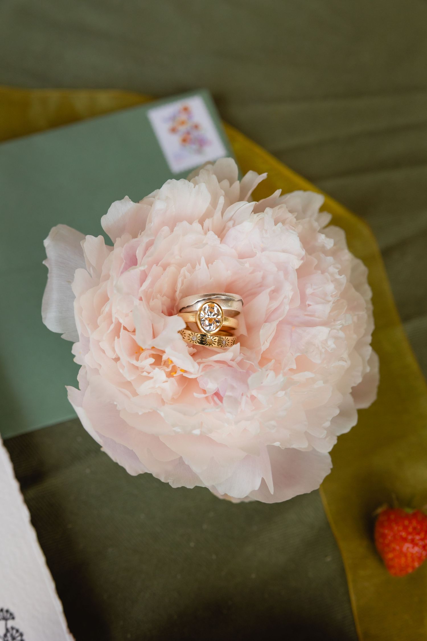 A delicate pink peony flower with wedding rings nestled in its soft petals against a dark background.