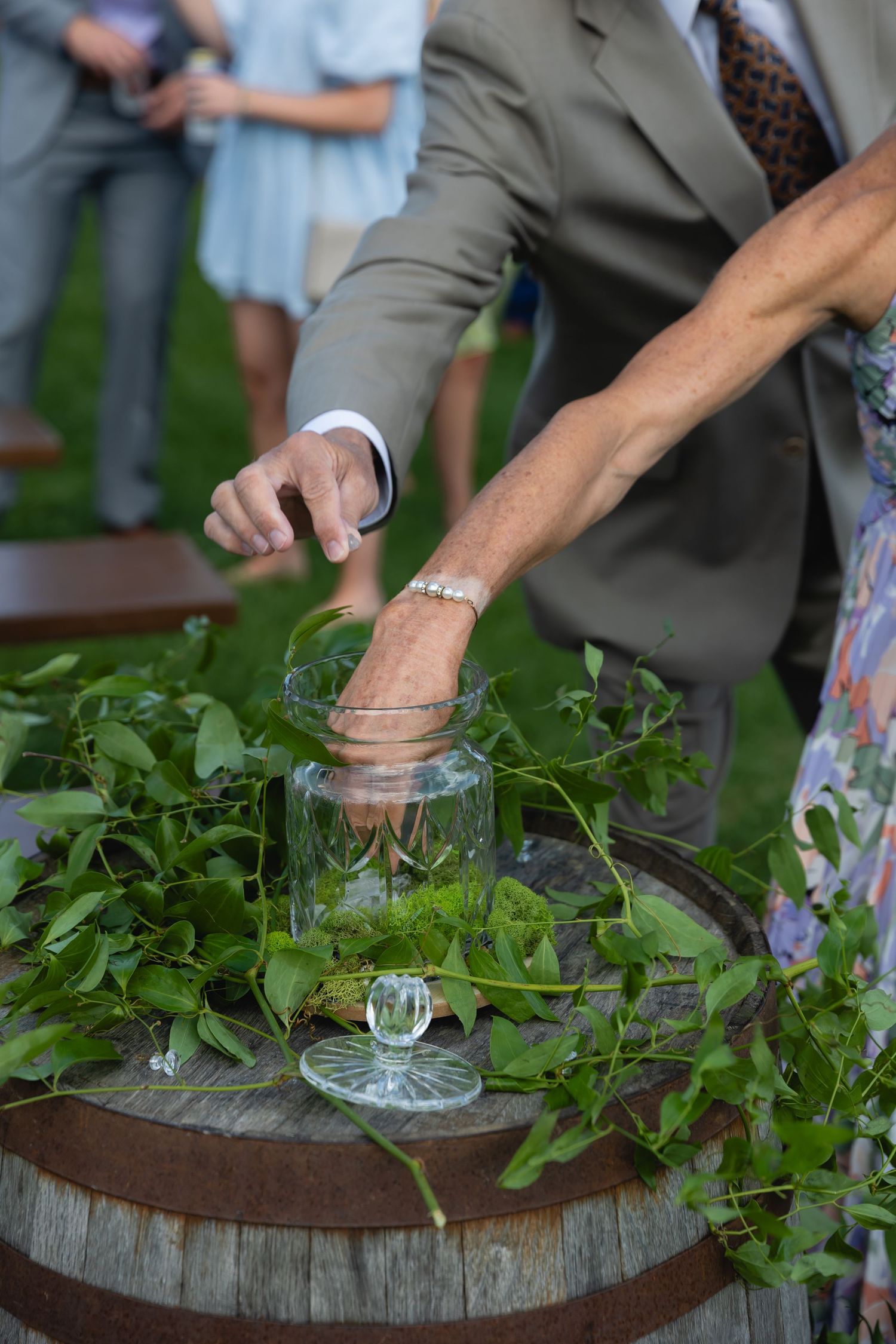 Hands placing fresh herbs into a glass jar on a rustic wooden barrel at an outdoor event.