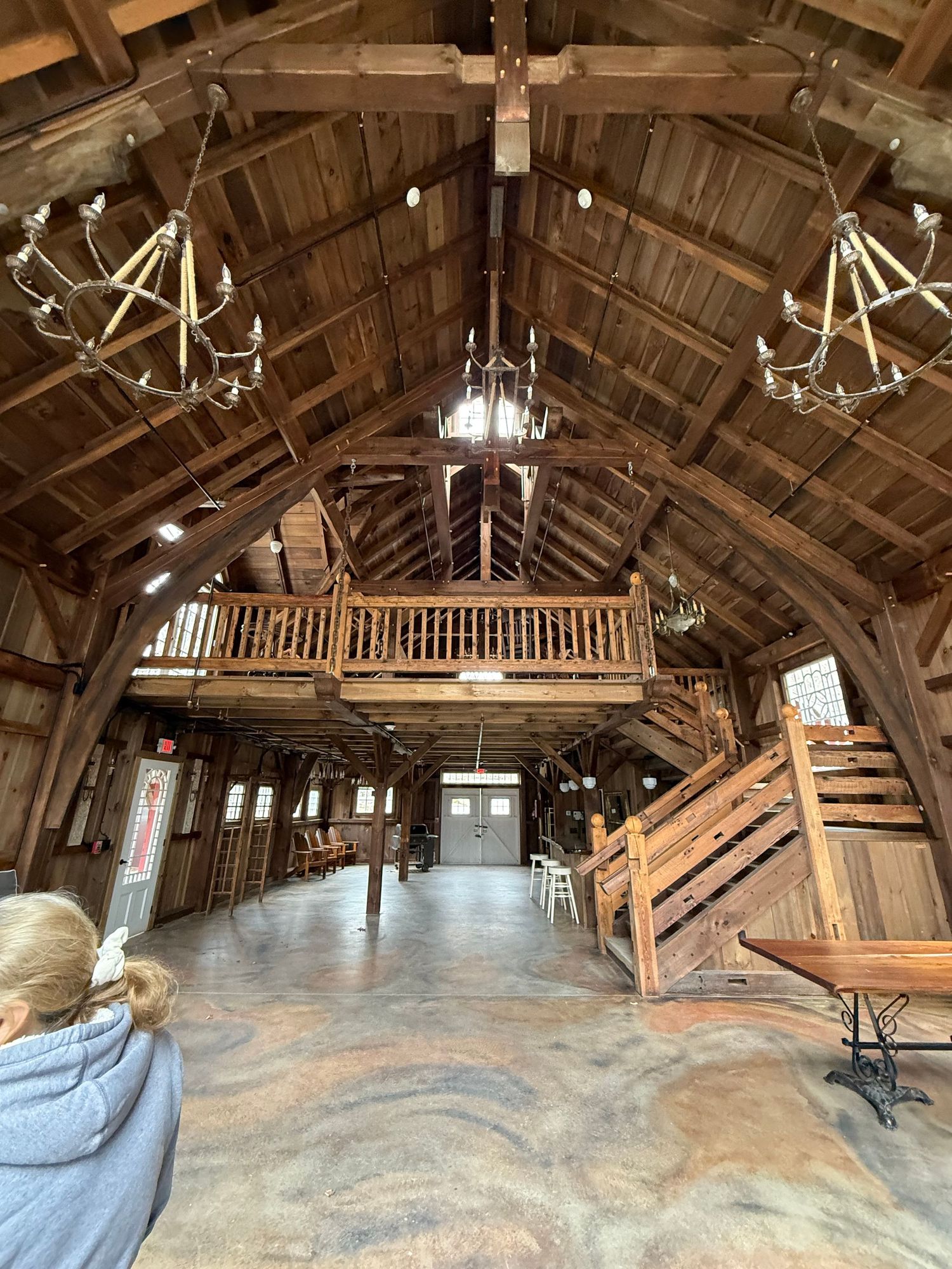 Rustic wooden barn loft interior with exposed beams, balcony railings, and chandeliers showing raw architectural features.
