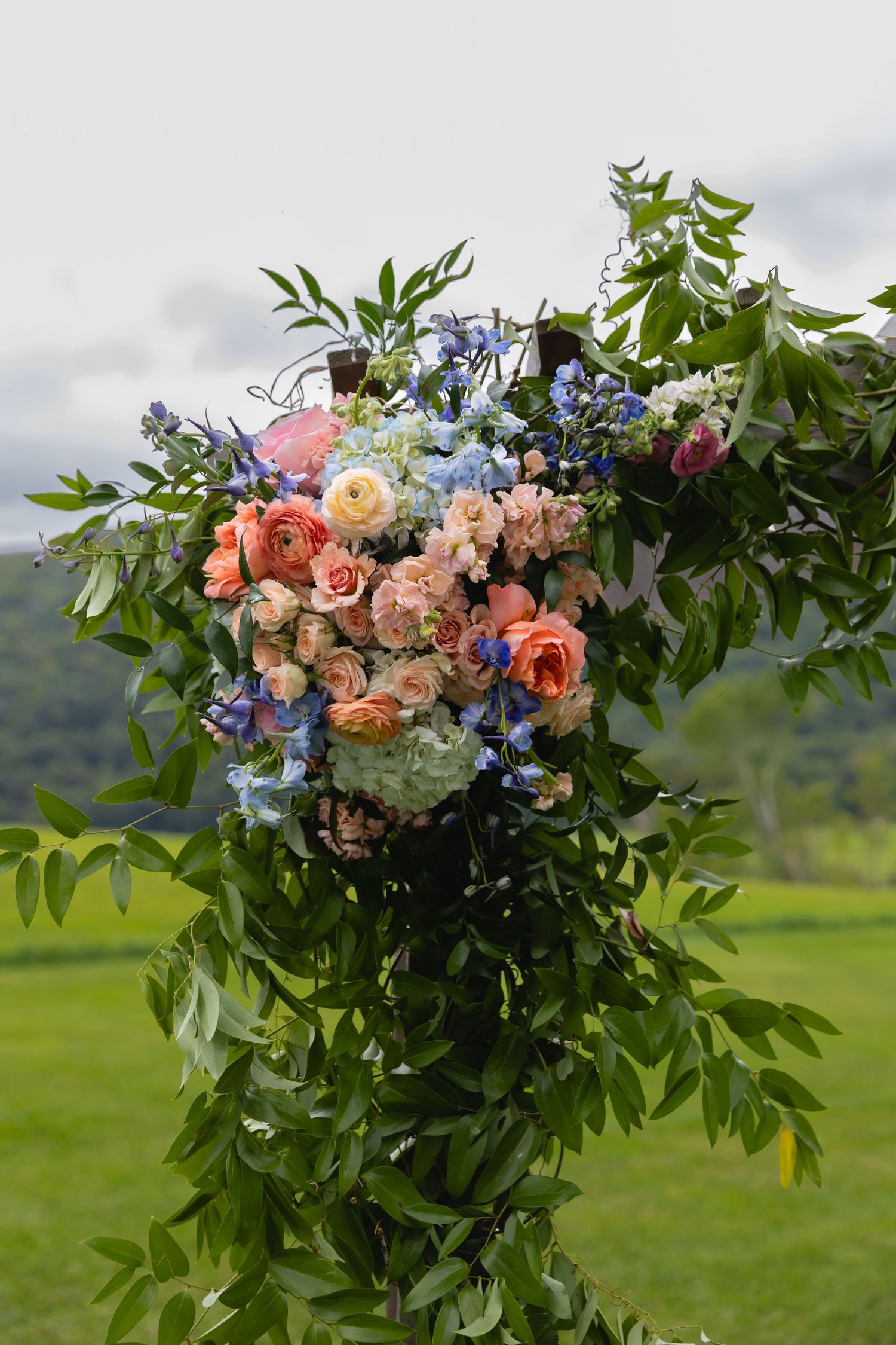 A lush floral arrangement with coral roses, blue delphinium and greenery displayed outdoors against a mountain landscape.