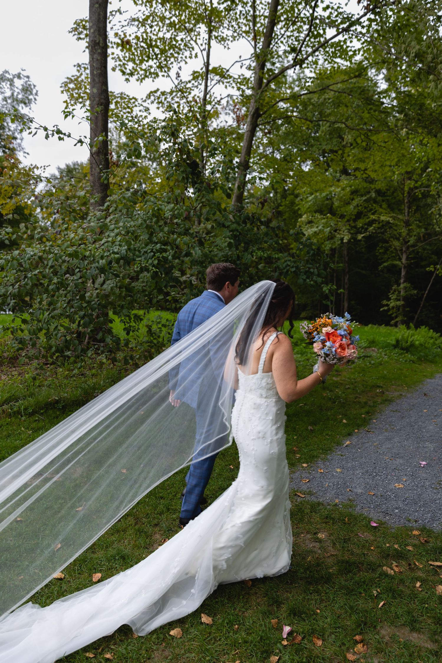 A couple poses outdoors with a flowing veil catching the breeze as the person in white holds a bright floral bouquet.