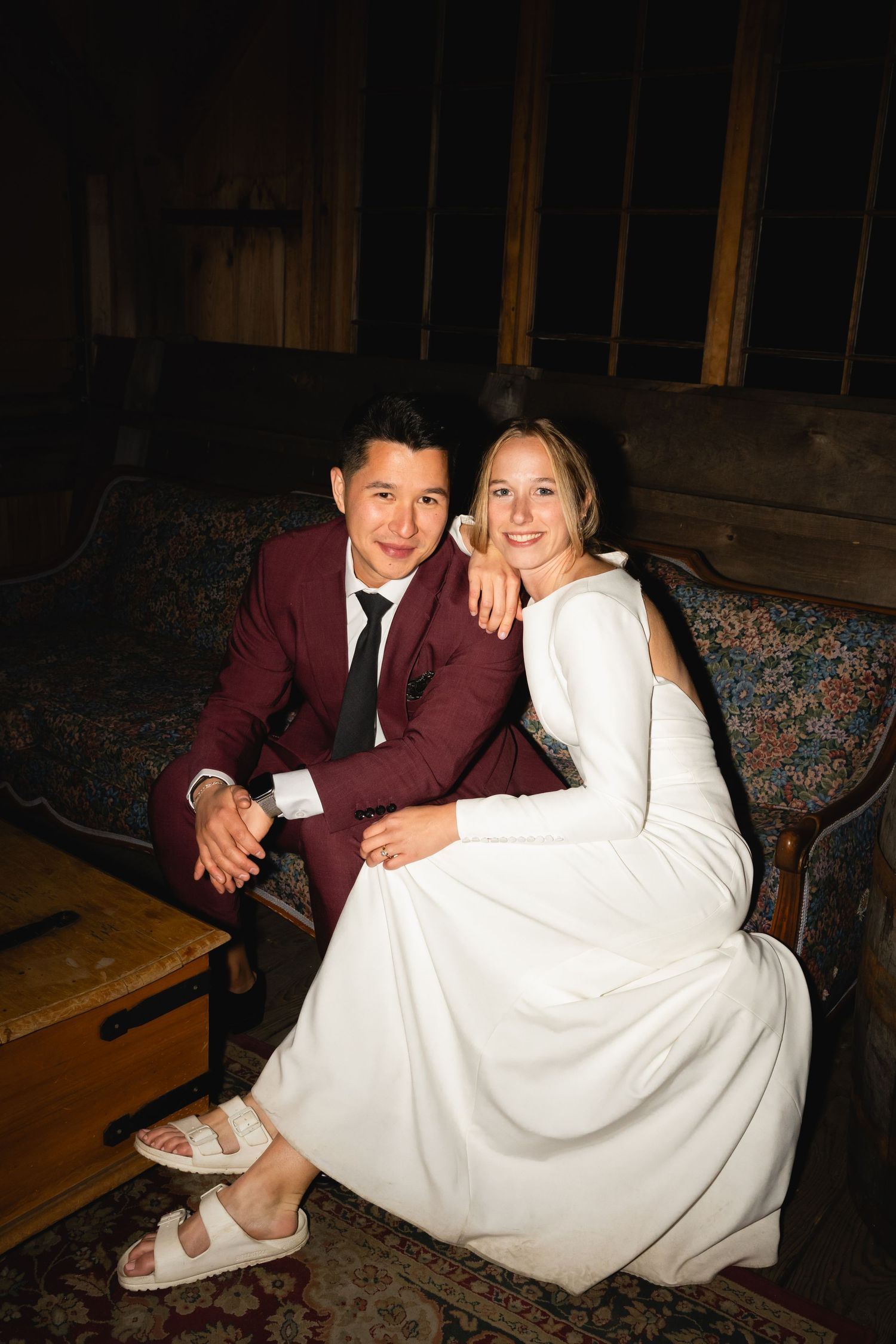 A couple sits together on an antique couch at their wedding, with the bride wearing white and groom in burgundy suit.