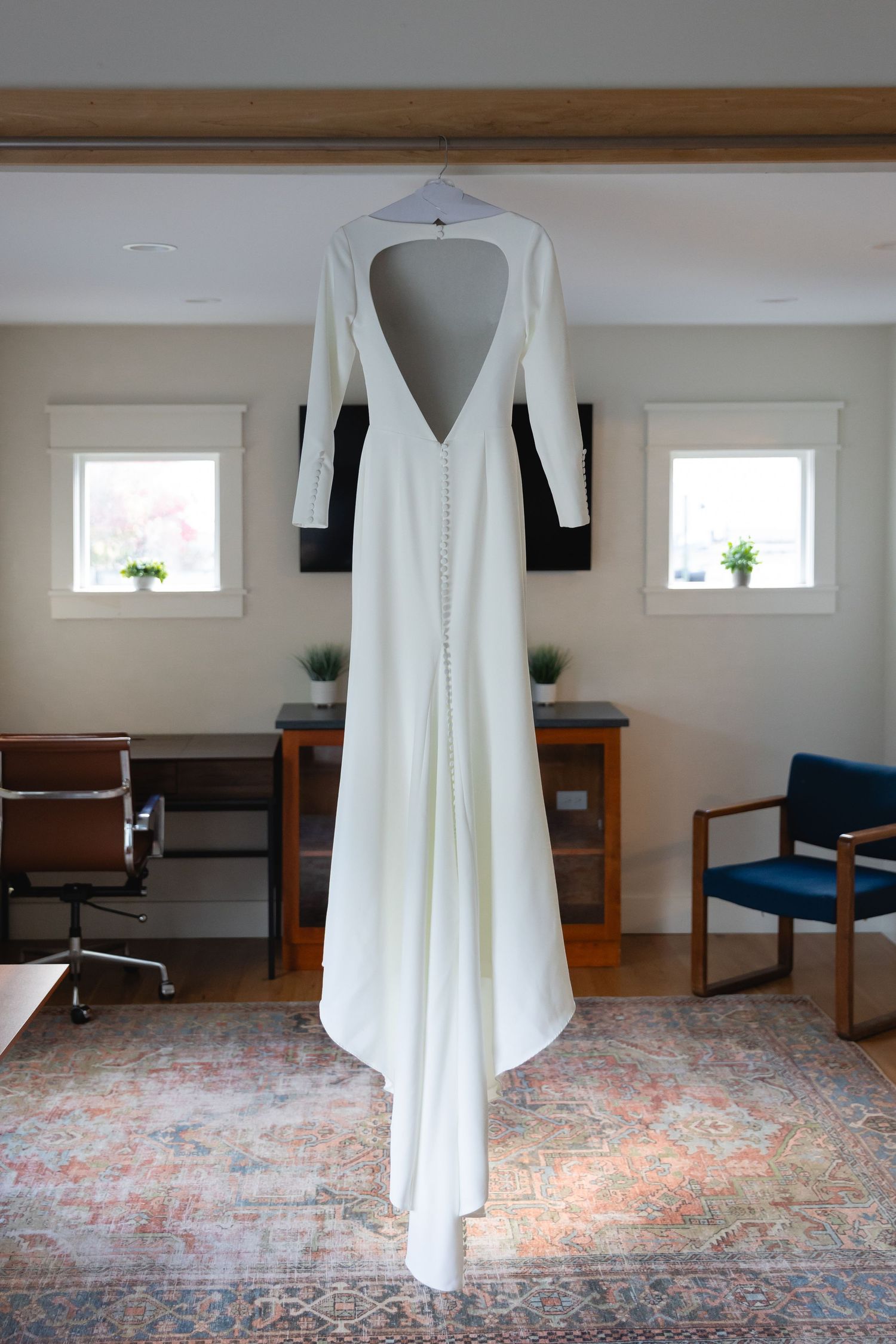 A modern white wedding dress with long sleeves and deep V-back hangs elegantly against a white wall between two small windows.