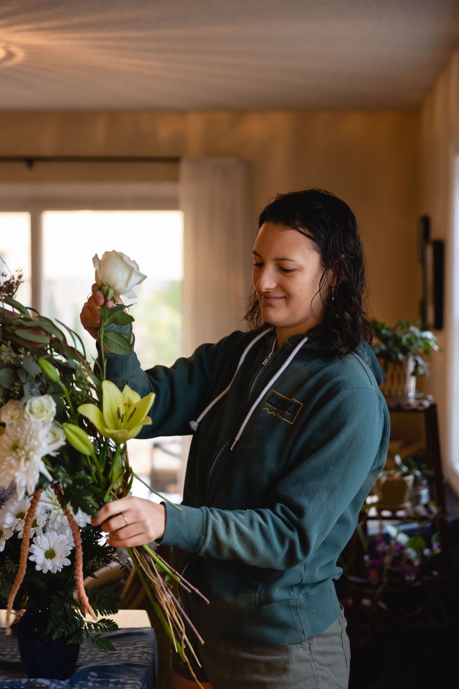 A florist arranges white roses and lilies at a sunlit workstation while wearing a teal sweatshirt.