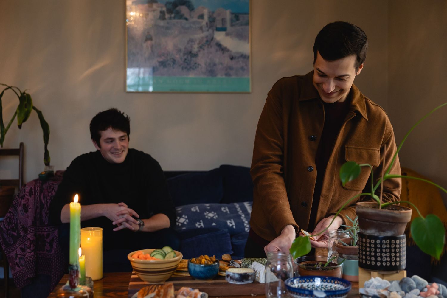 Two people cooking and preparing food at a dining table with drinks and kitchen items in a cozy home setting.