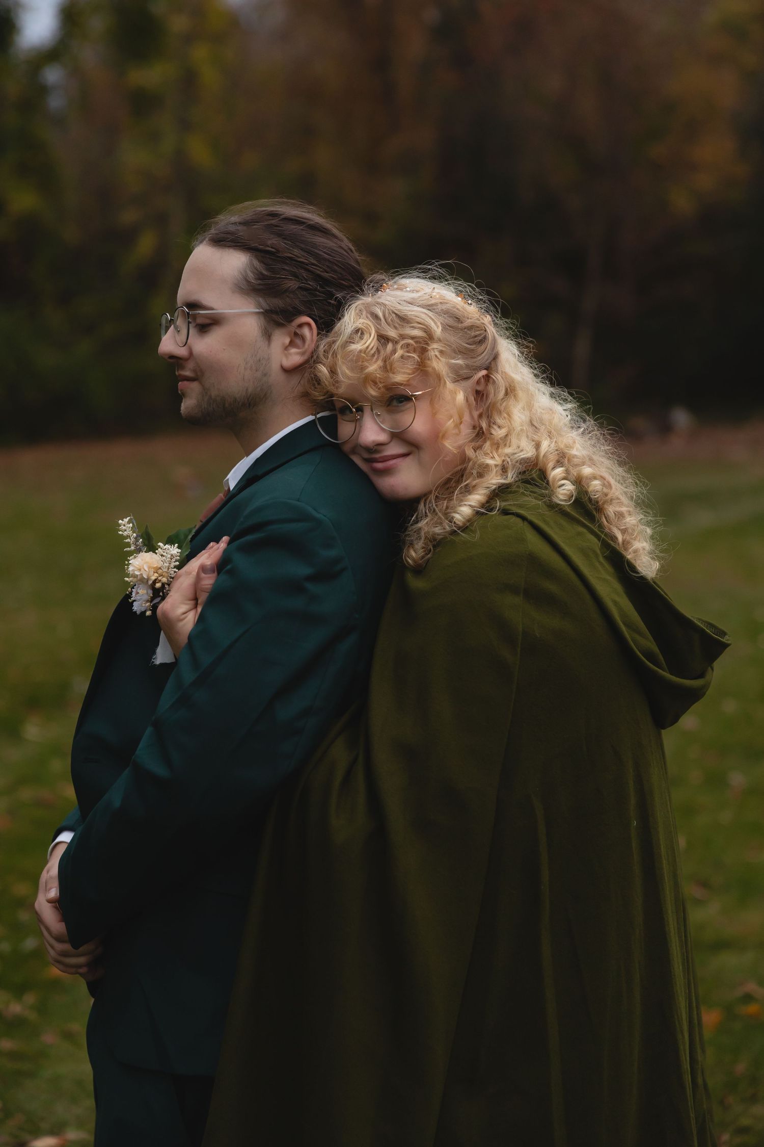Couple in green cape and dark suit embrace during autumn outdoor photo session.