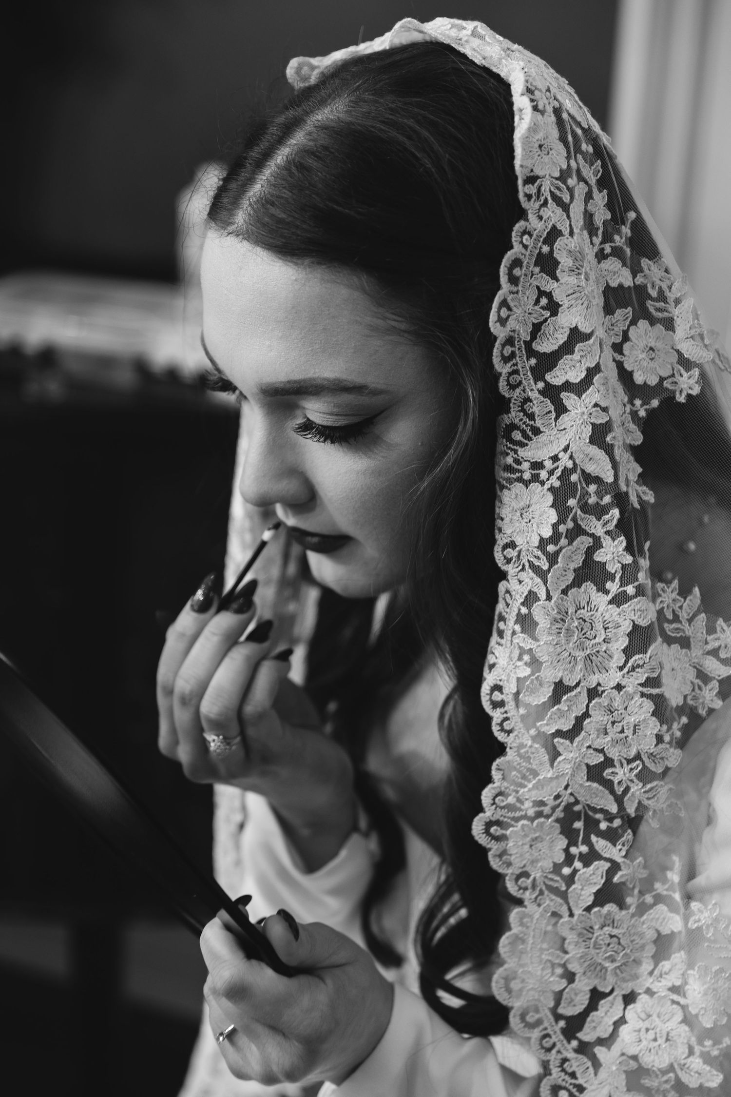 A black and white portrait of a bride wearing a delicate lace veil while applying lipstick.