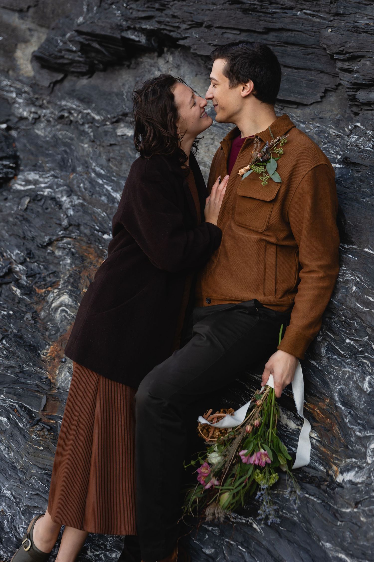 Romantic couple embraces against dark rocky background, person in brown coat holding fresh floral bouquet.