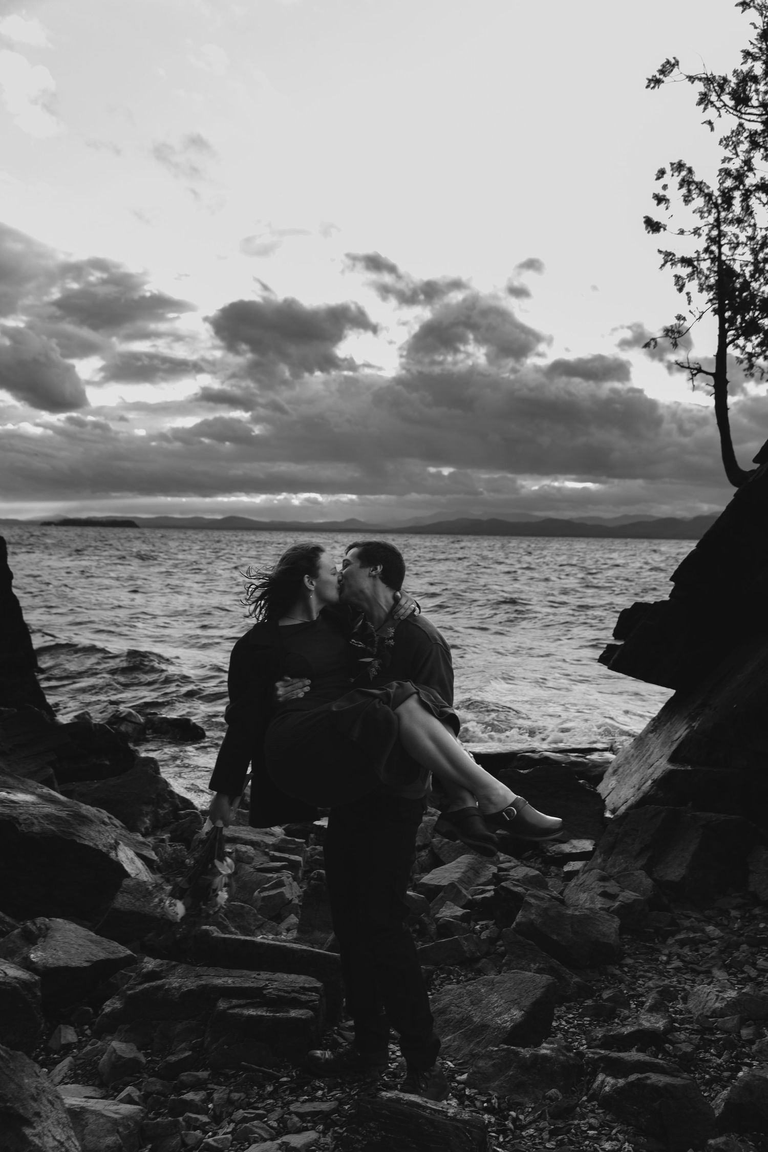 Romantic silhouette of a couple sharing an intimate moment on rocky shore with dramatic cloudy sunset over the water.