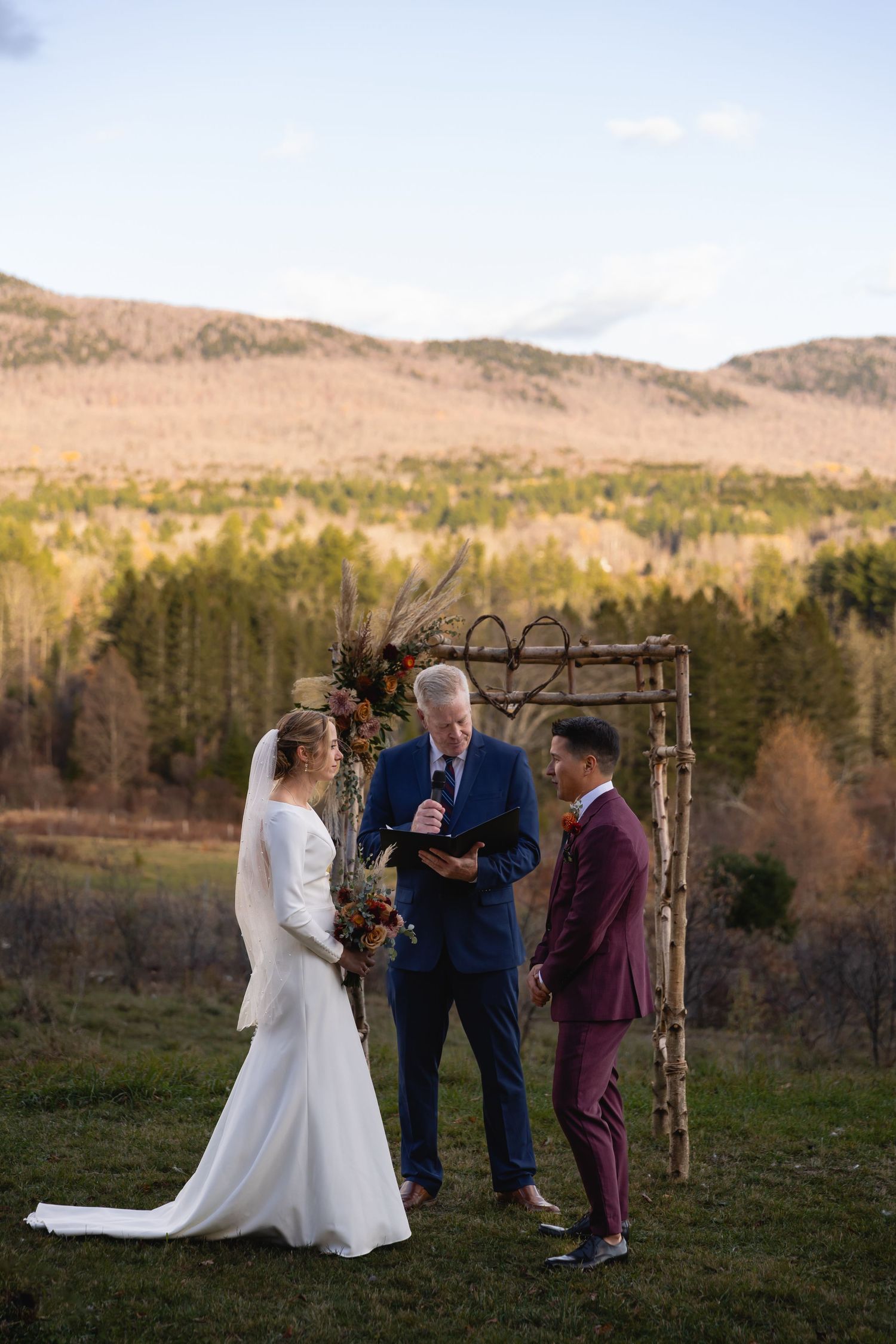 A wedding ceremony takes place under a rustic wooden arch with mountains and forest in the scenic background.