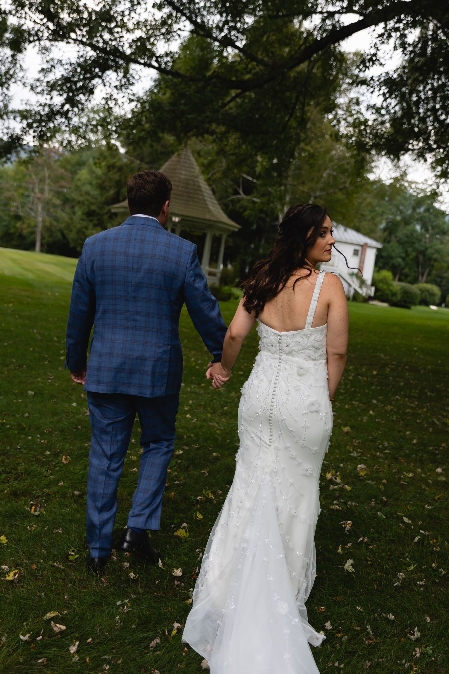 A couple in wedding attire walk hand in hand across a green lawn with a rustic chapel in the background.