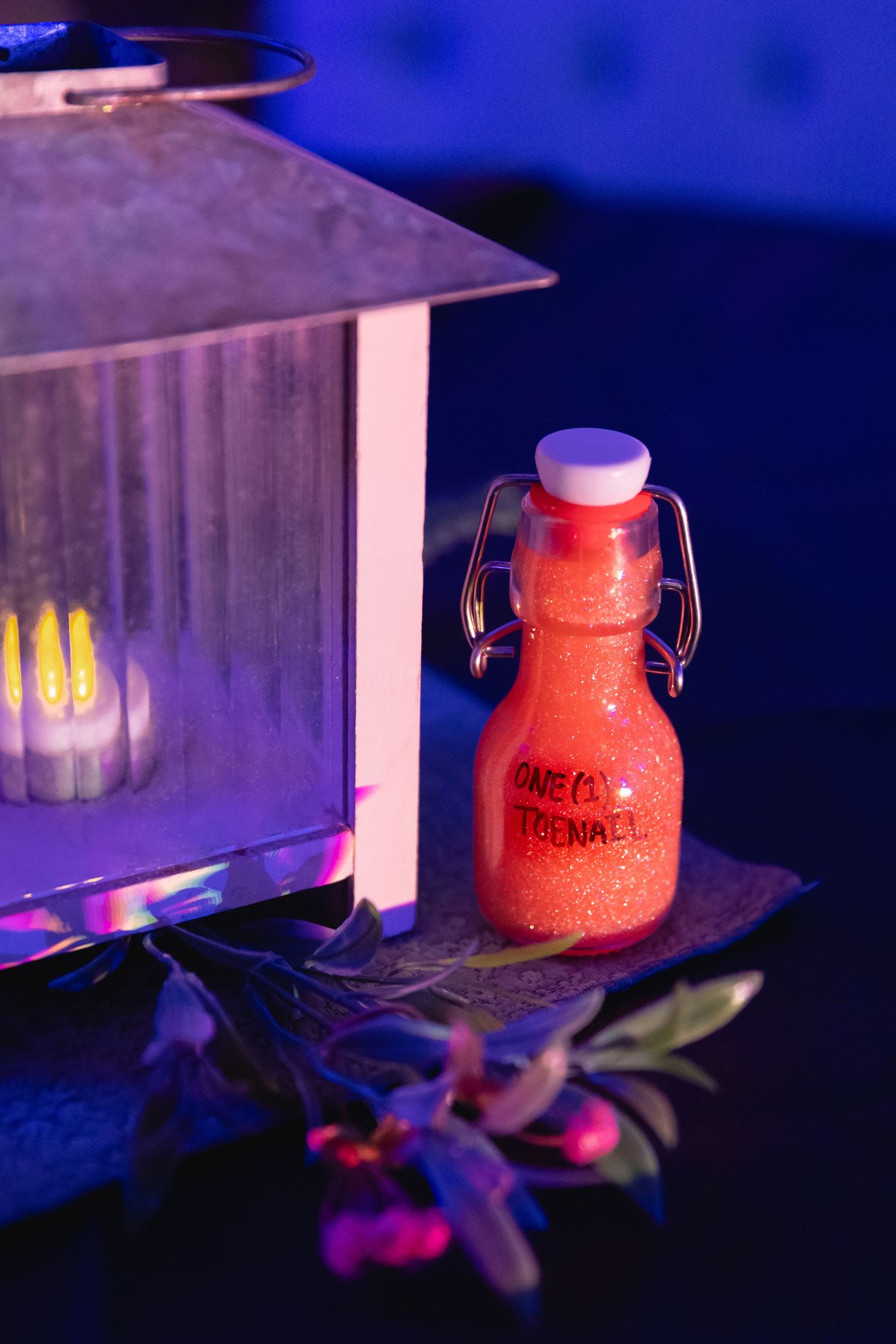 Small red glitter potion bottle labeled as wedding favor displayed beside a metal lantern with candle.
