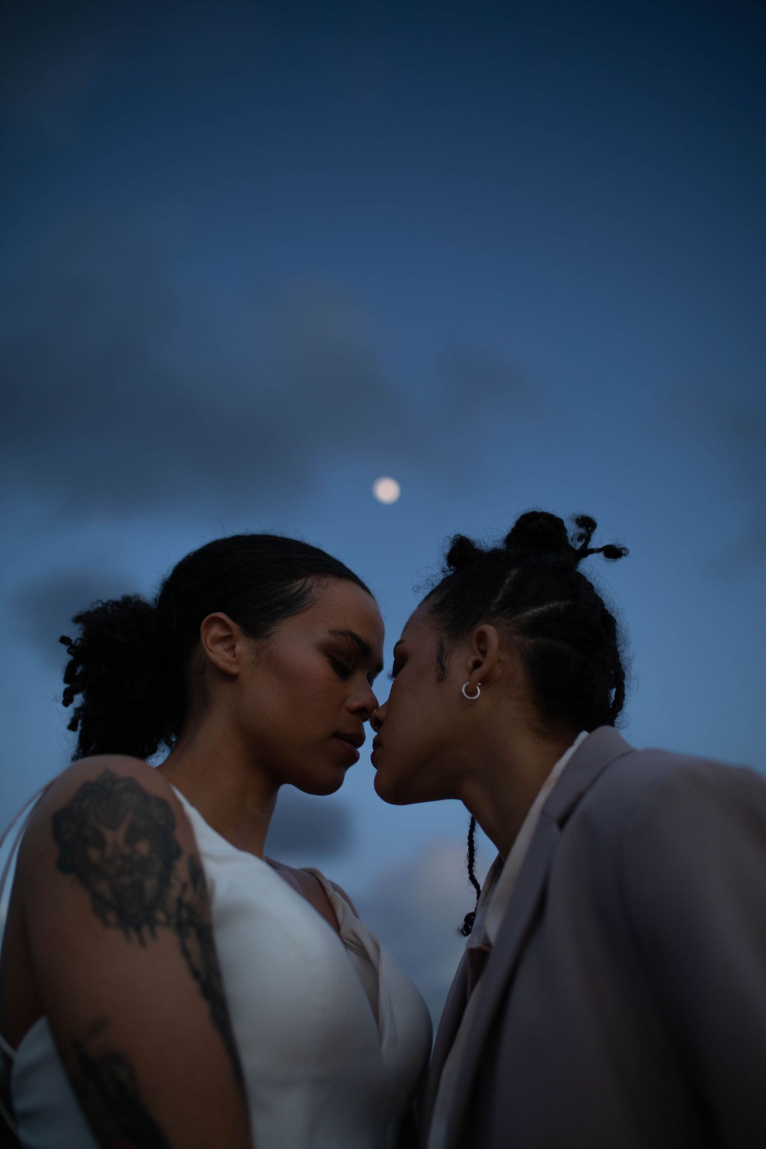 Two brides share intimate kiss under moonlit blue sky at evening wedding ceremony.