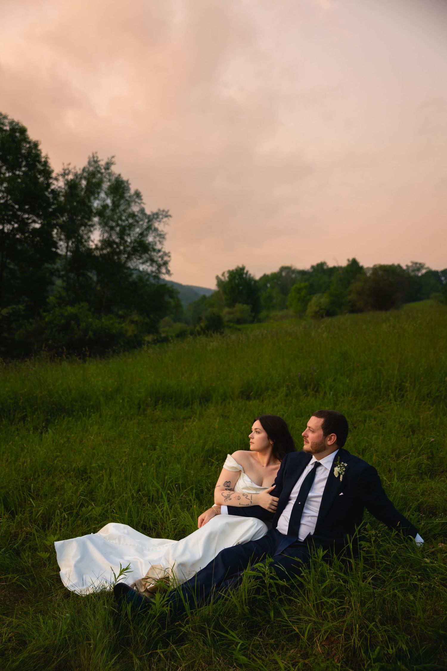 Bride and groom sitting in green meadow at sunset with pink cloudy sky above.