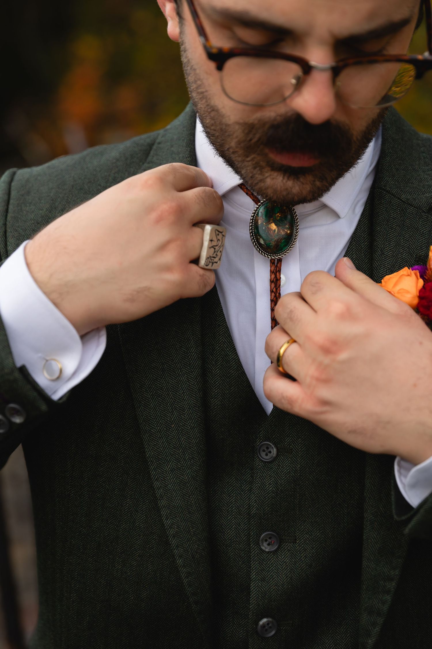 Groom in green suit adjusting bolo tie with floral boutonniere in autumn setting.