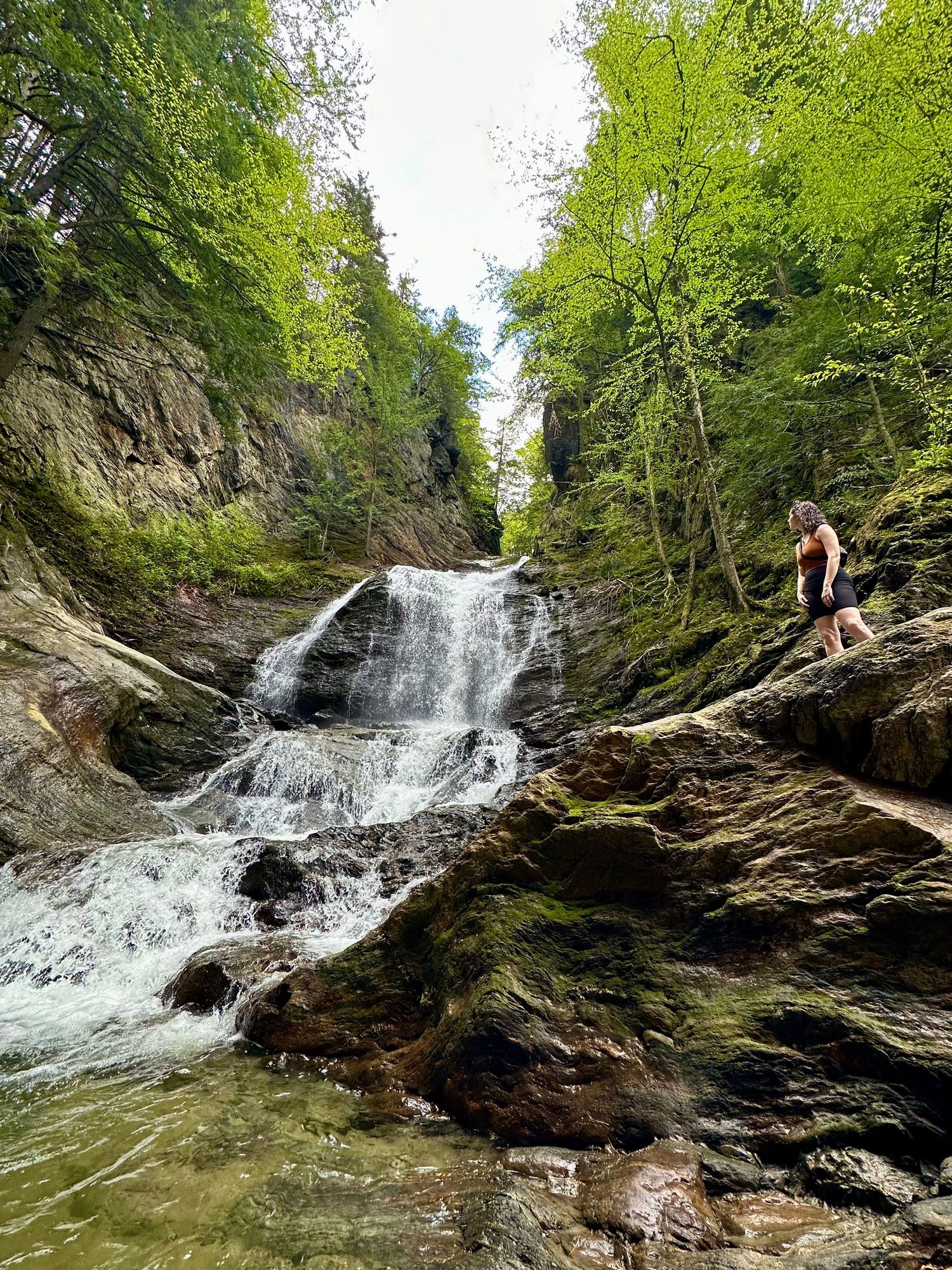 Woman standing on mossy rocks beside a cascading waterfall surrounded by lush green forest trees.