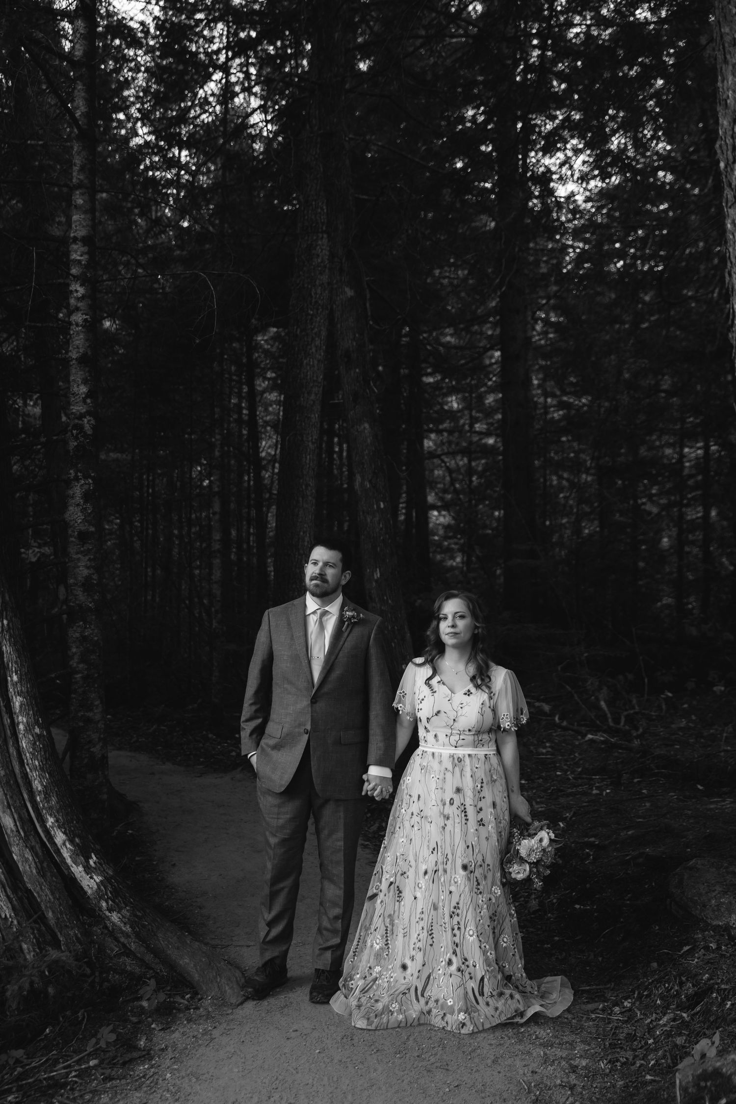 A couple in wedding attire stands on a forest path surrounded by tall trees in a black and white photograph.
