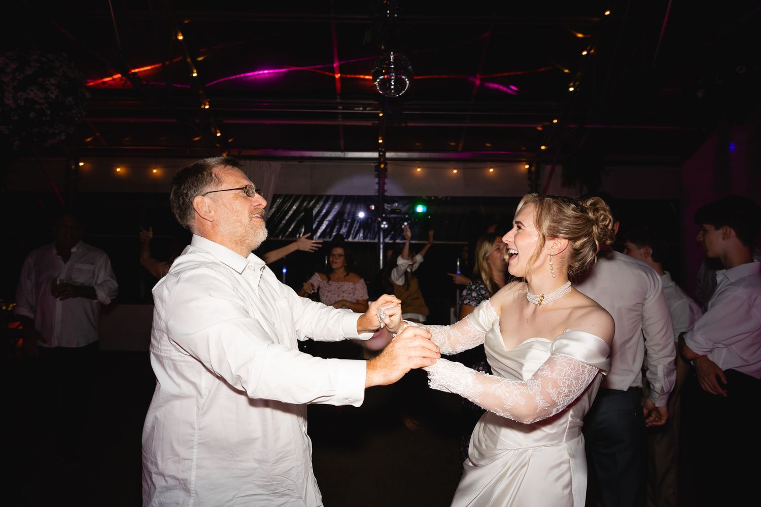 A couple dances together at a dimly lit venue with colorful lighting in the background.
