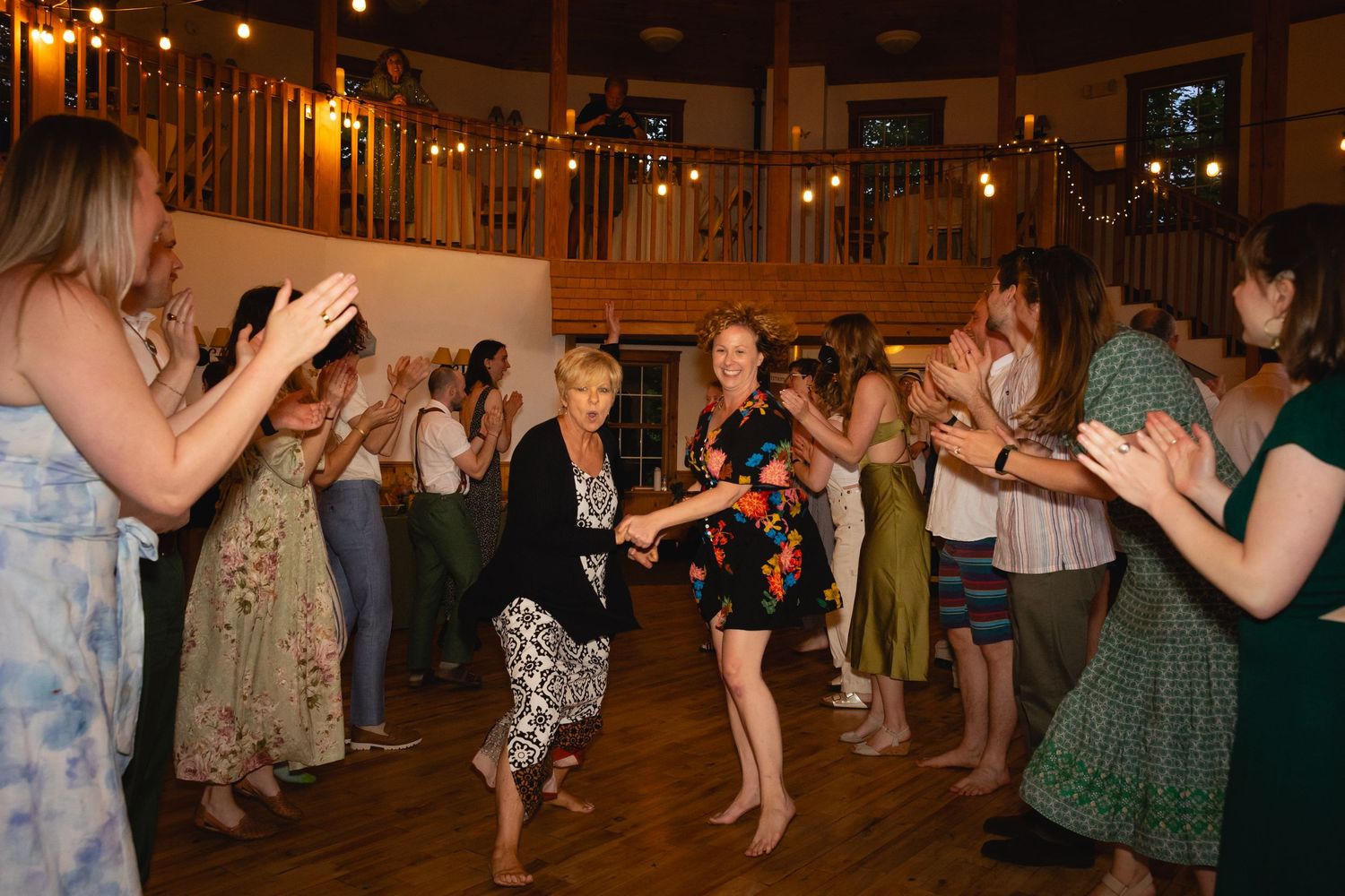 People dancing and celebrating in a warmly lit wooden dance hall with string lights overhead.
