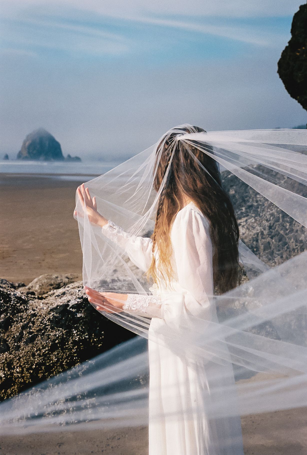 A flowing white wedding dress and veil catch the ocean breeze on a misty beach with rocky cliffs in the background.