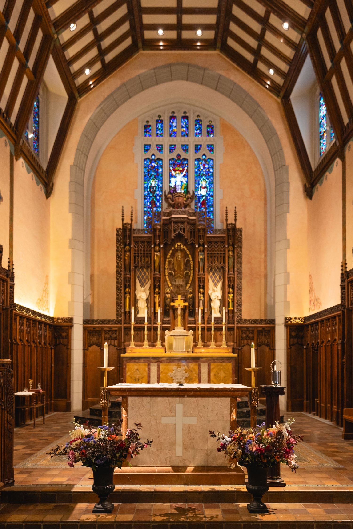 Gothic church altar with wooden beams, stained glass windows, and ornate decorative elements illuminated by warm lighting.