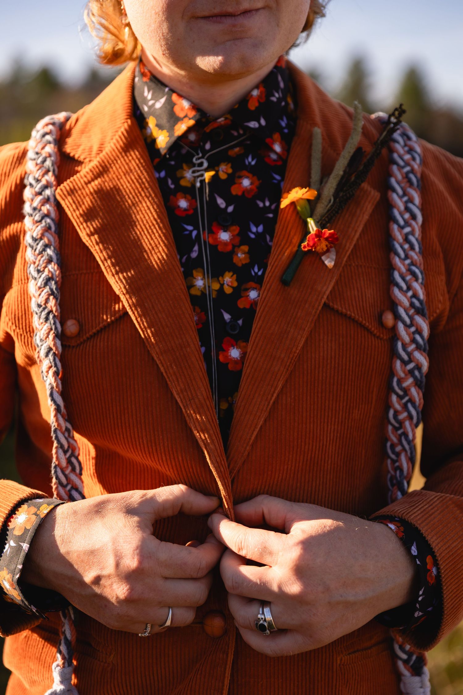 Close-up of someone wearing an orange corduroy blazer with floral shirt, braided suspenders and boutonniere.