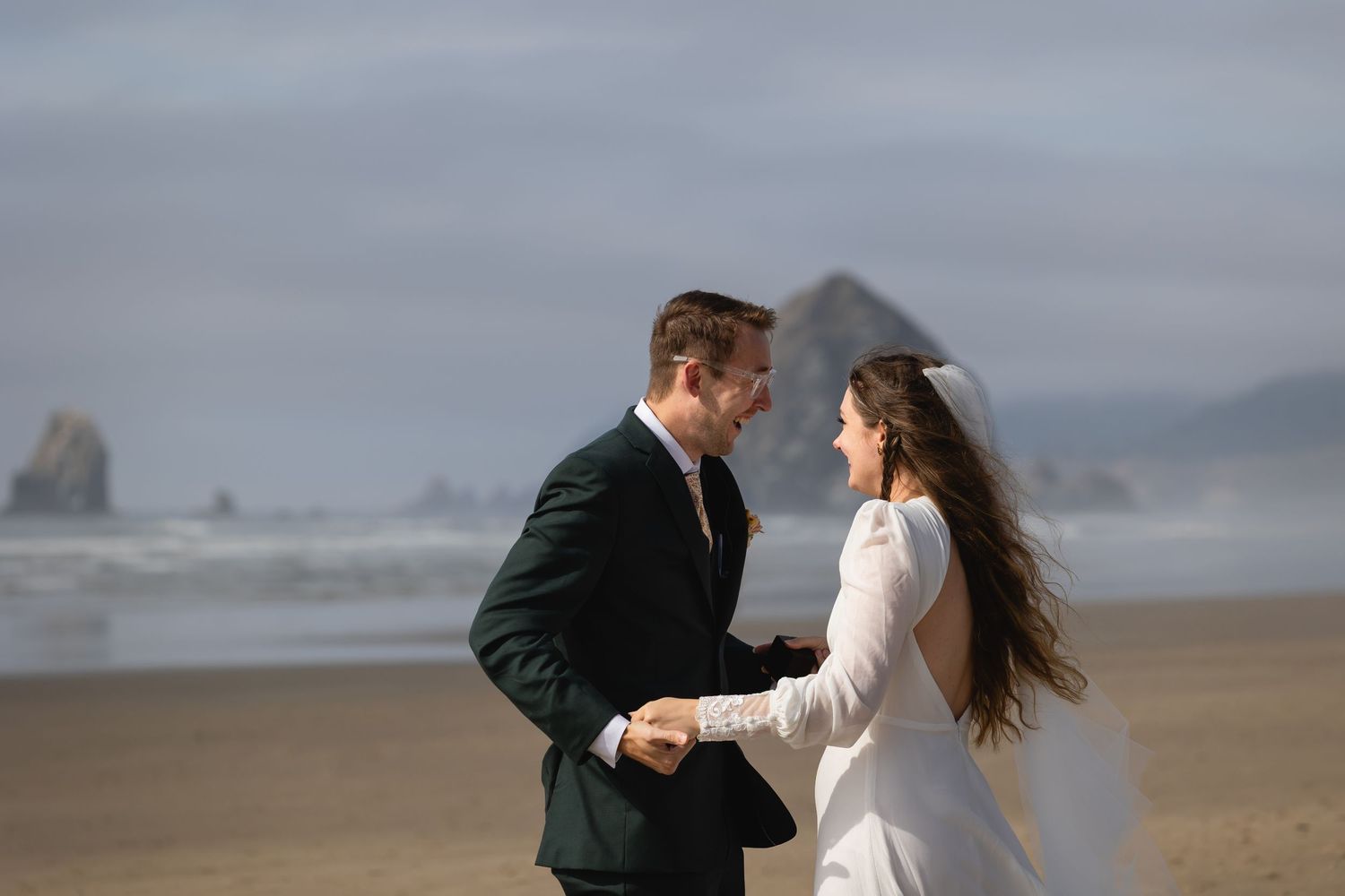 A couple in wedding attire stands together on a windy beach with dramatic cloudy skies in the background.