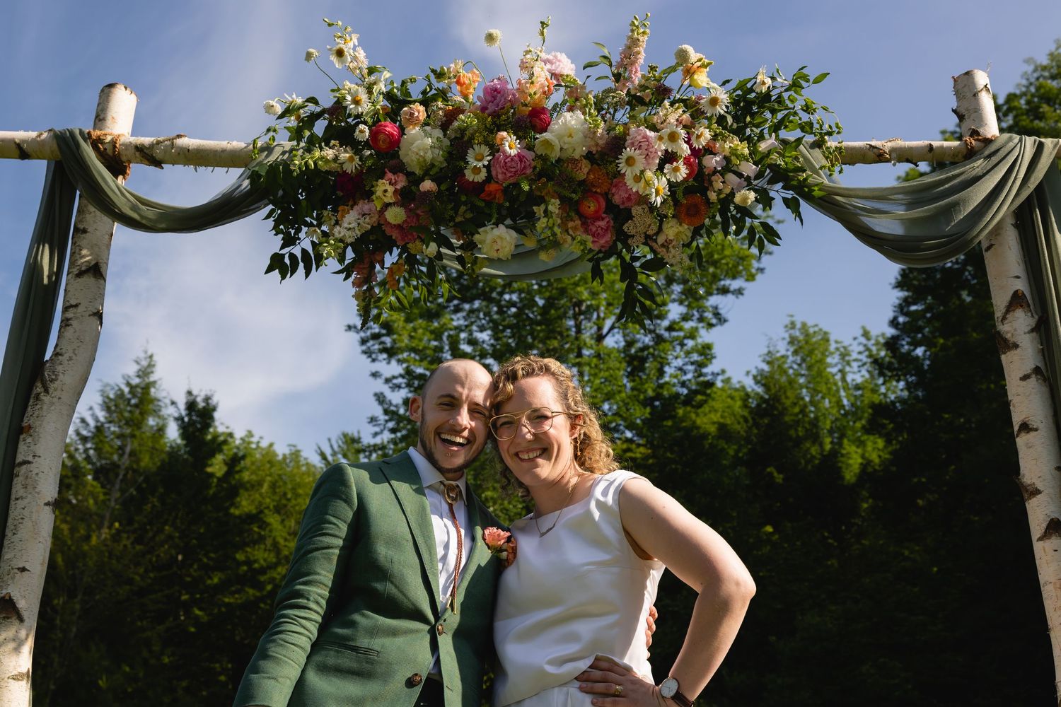 A rustic wooden arch decorated with lush floral arrangements featuring pink and white flowers against a blue sky.