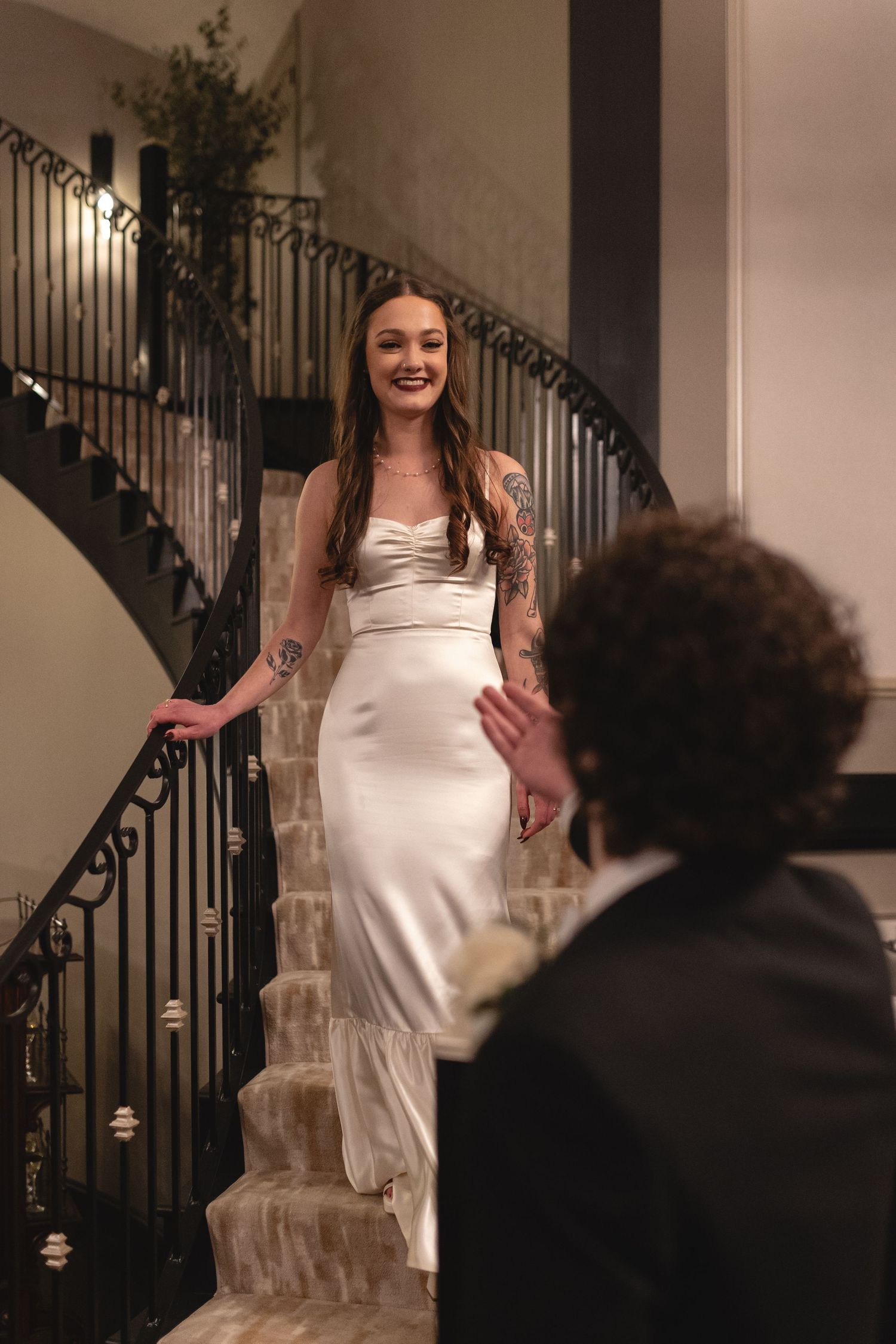 A bride in an elegant silk wedding dress poses on a curved staircase with illuminated railings.