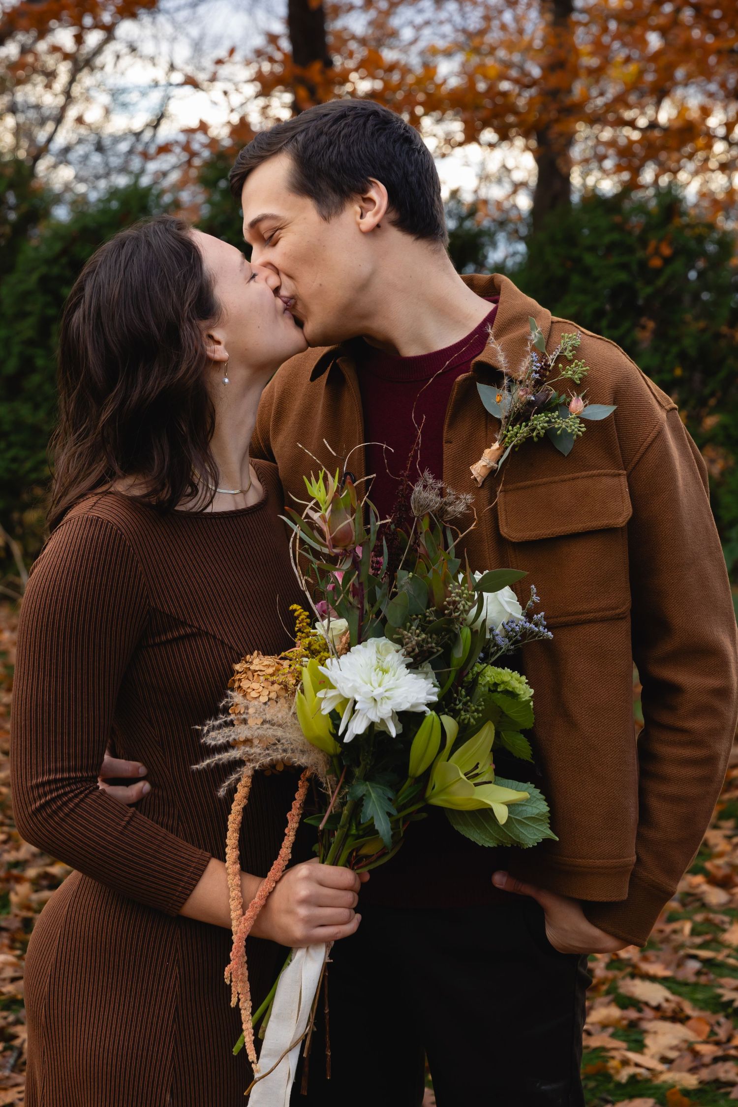 A romantic autumn scene shows a couple sharing a kiss while holding a rustic flower bouquet in a fall-colored setting.