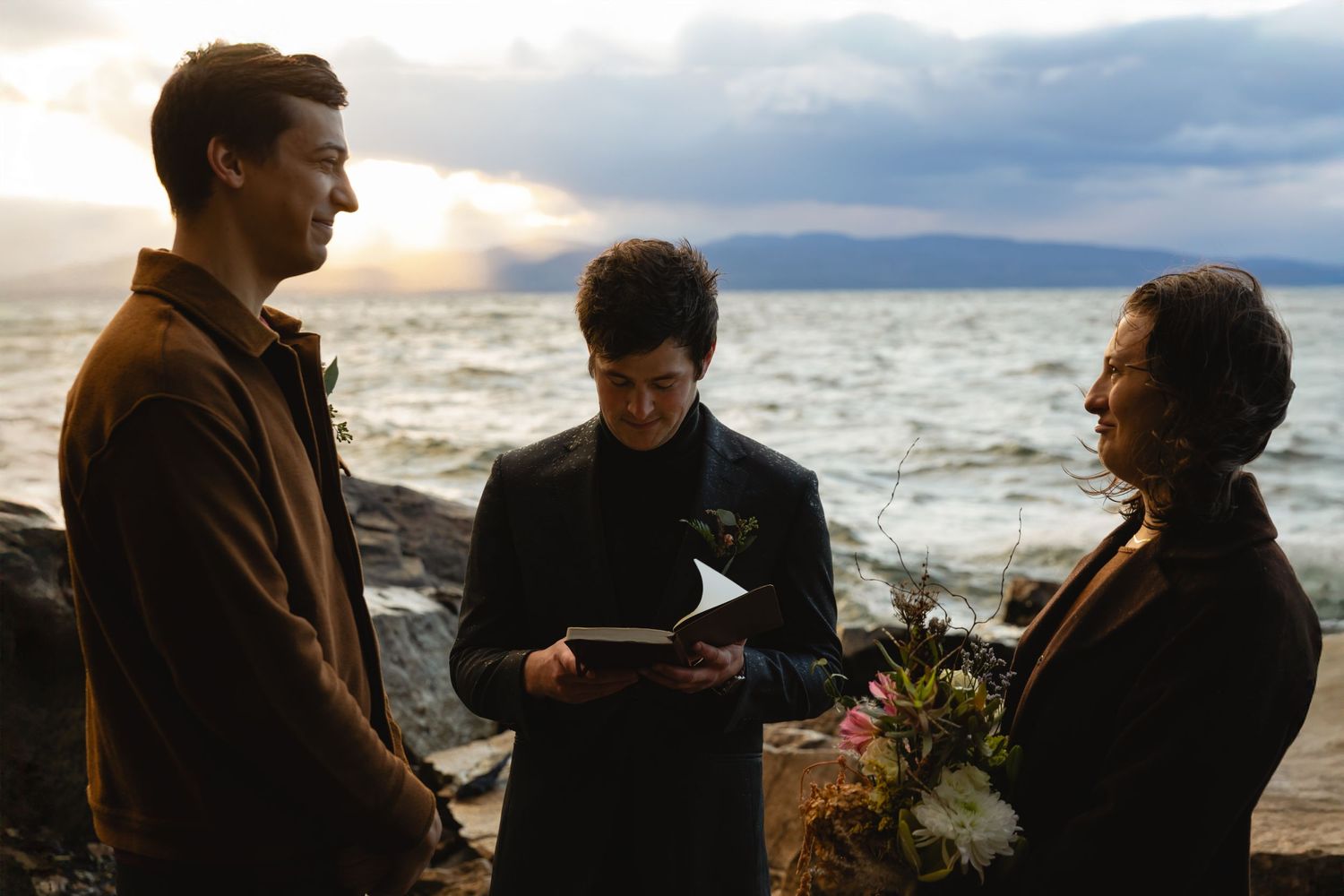 Three people gather for a seaside ceremony at sunset with mountains in the background.
