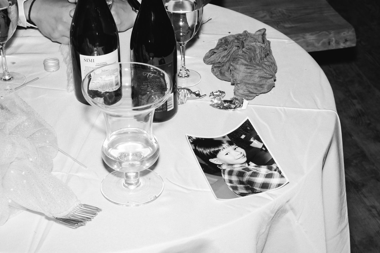 A black and white photograph shows wine bottles, glasses, and a photograph on a table with white tablecloth.