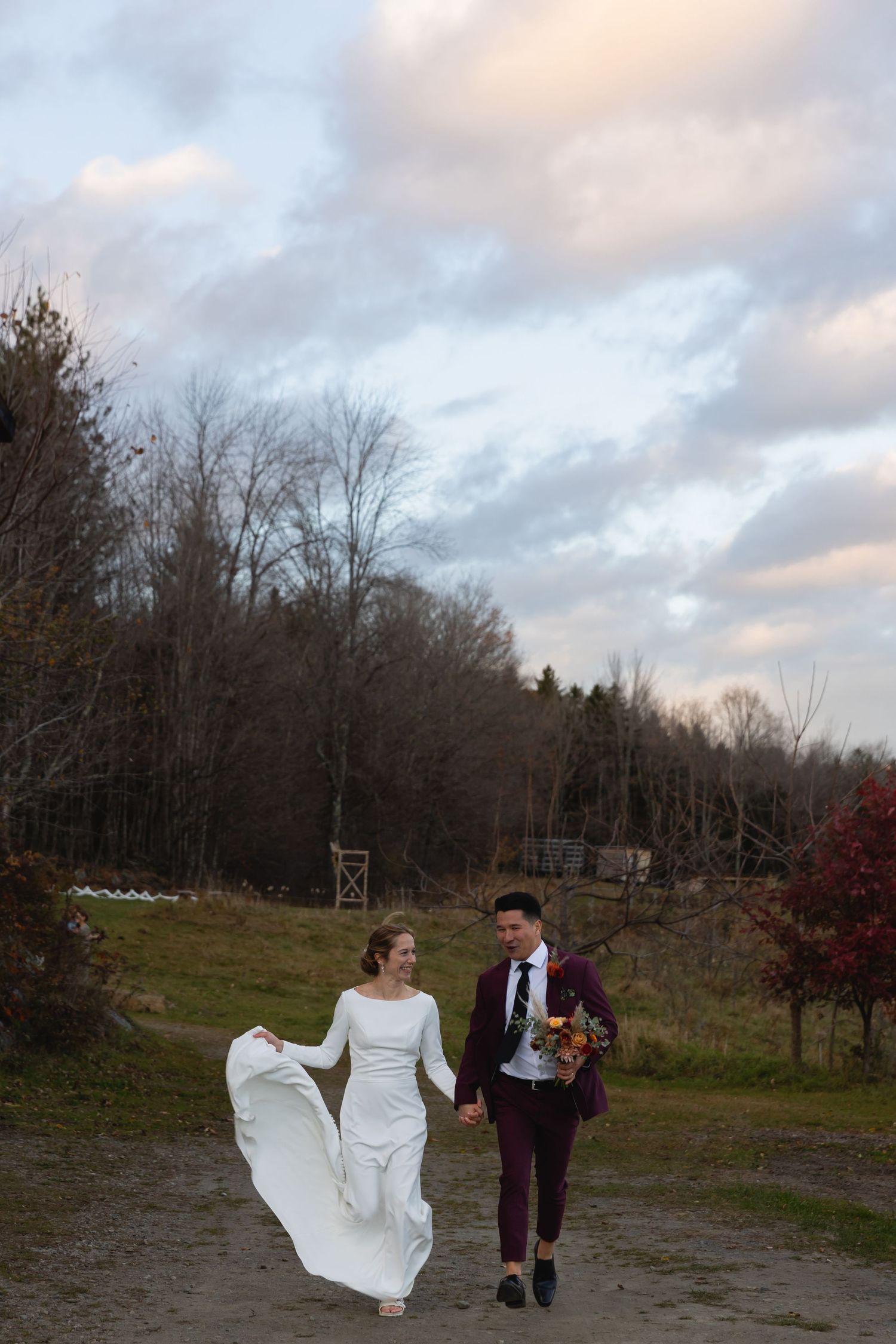 A couple walks along a dirt path in autumn, with the bride's white dress flowing in the wind against a cloudy sky backdrop.