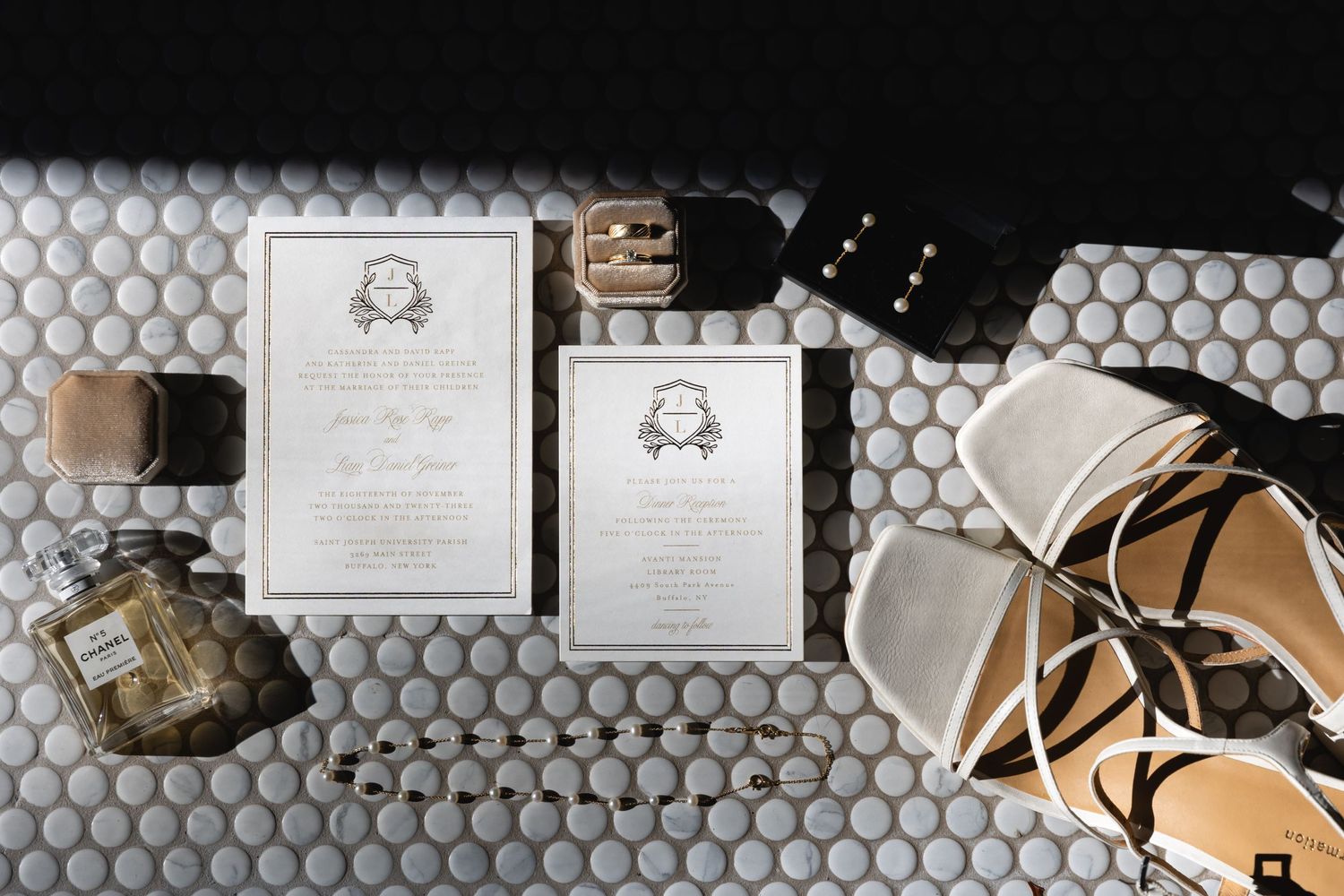 Wedding invitations with monogram design displayed on hexagonal tile floor alongside bridal shoes and perfume bottles.