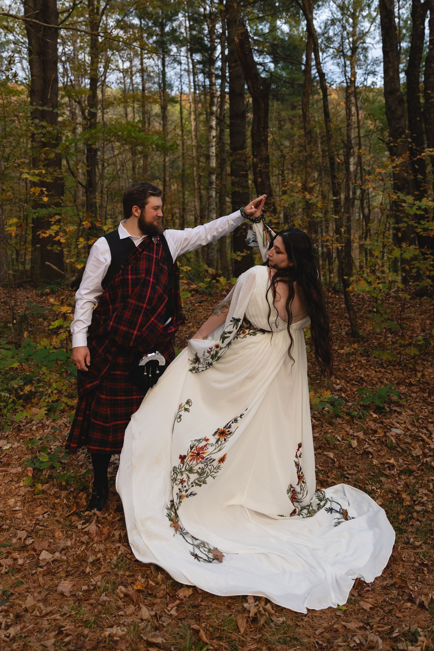 A couple dancing in autumn woods with traditional Scottish attire and an embroidered white wedding dress.