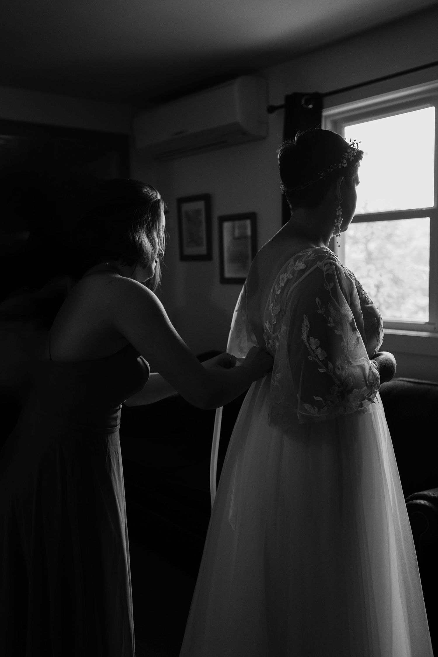 Black and white photo of a wedding dress being adjusted in a dimly lit room near a window.