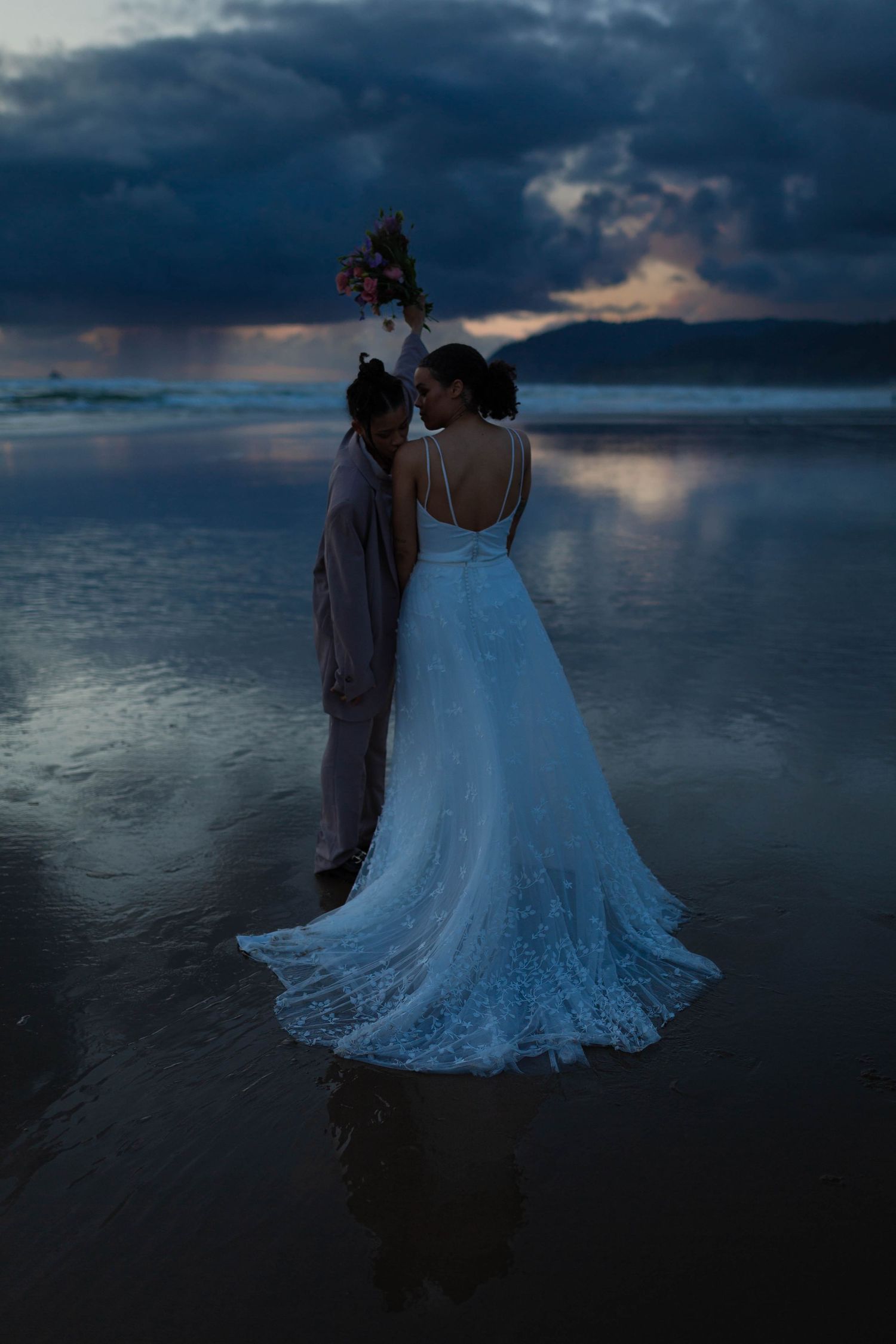 A couple shares an intimate moment on a beach at dusk with stormy skies and mountain backdrop.