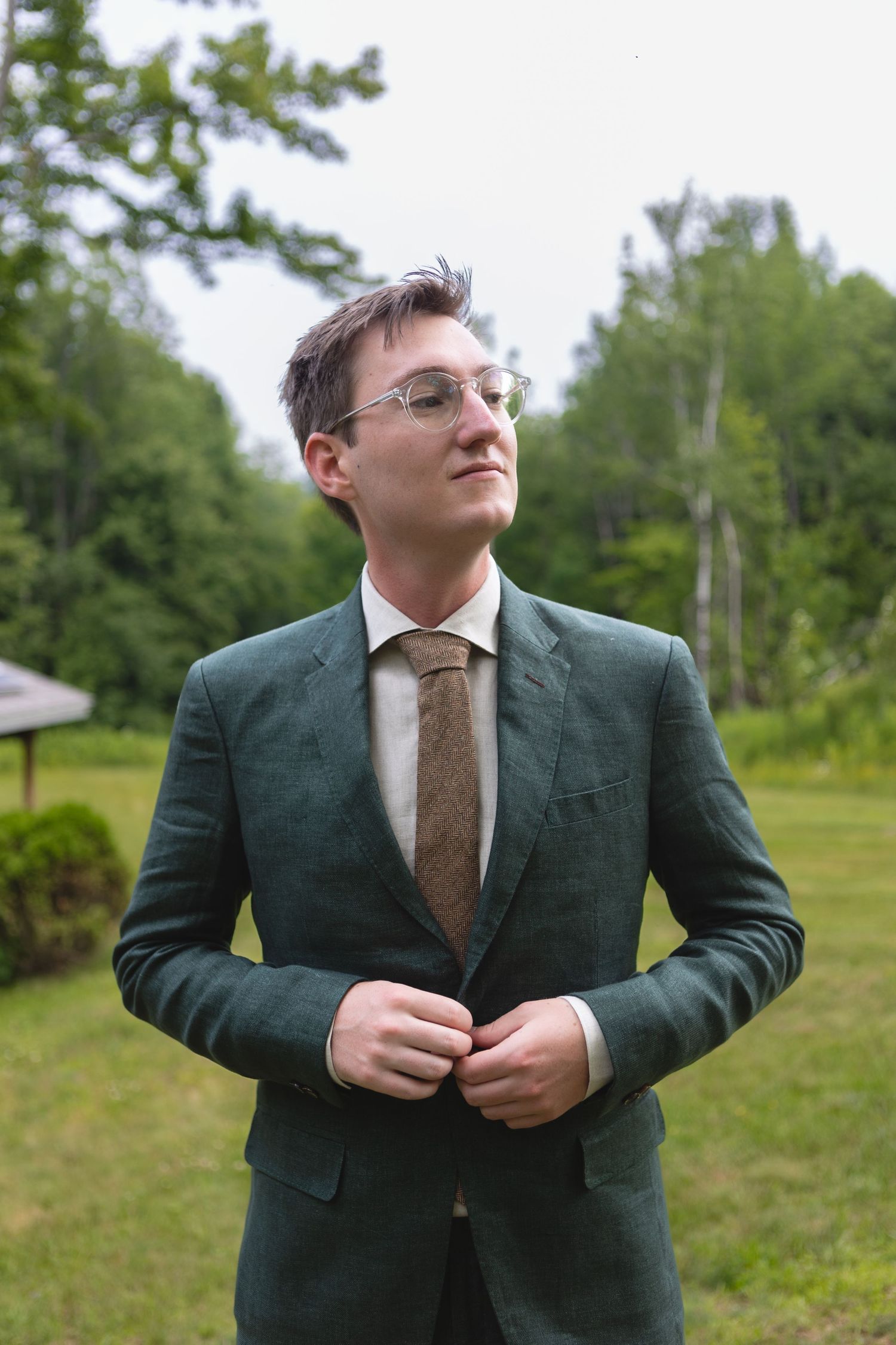 A groom in dark green suit and brown tie adjusts his jacket outdoors.